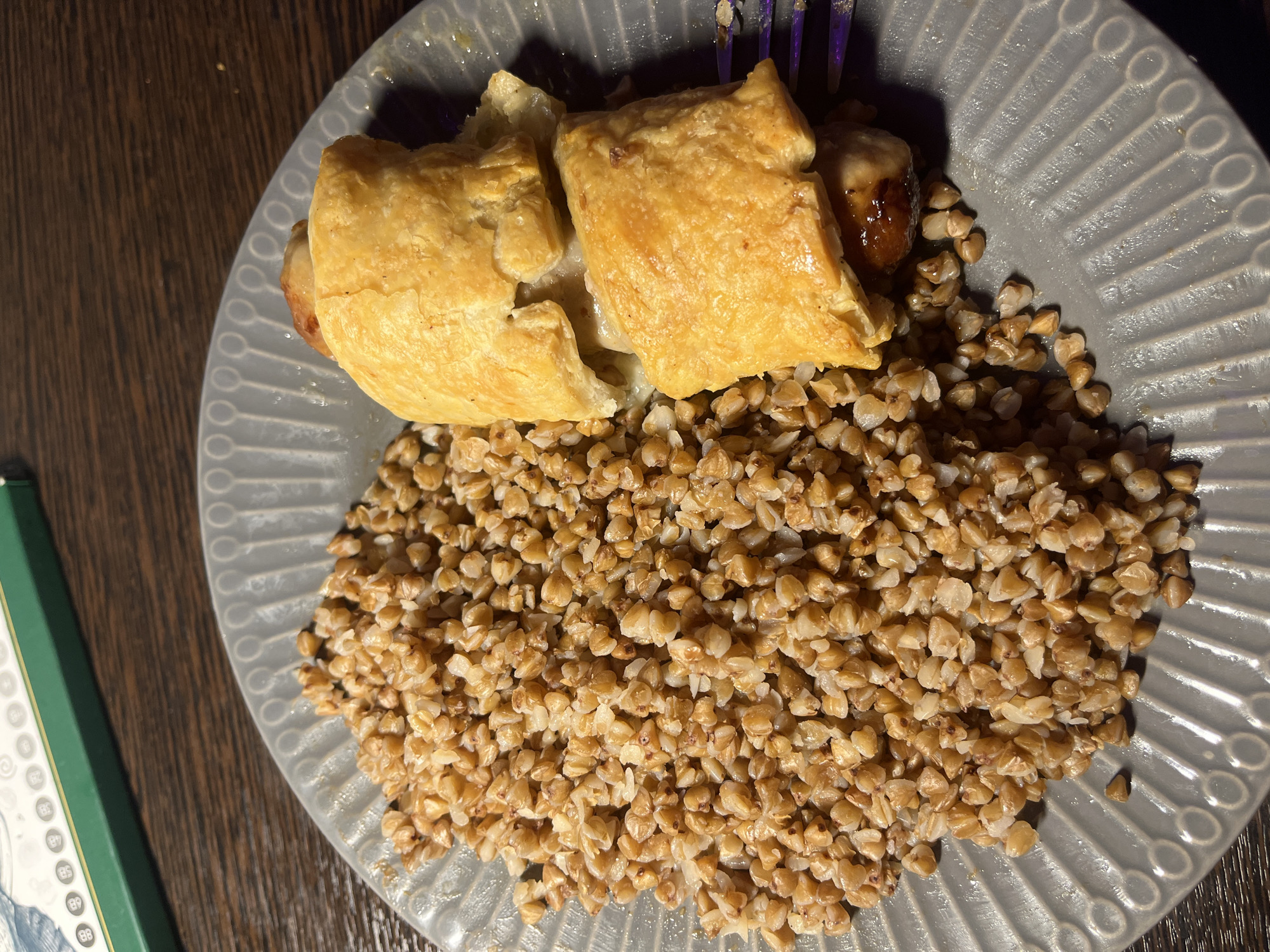 The image depicts a grey plate with a pastry and a portion of buckwheat kasha on a wooden surface. The pastry is golden brown and appears to be cut into two pieces. The kasha fills the lower portion of the plate. A blue and purple fork is visible near the pastry. A green box sits in the bottom-left corner of the image. It is impossible to know the location in Saint Petersburg, Russia without further information.