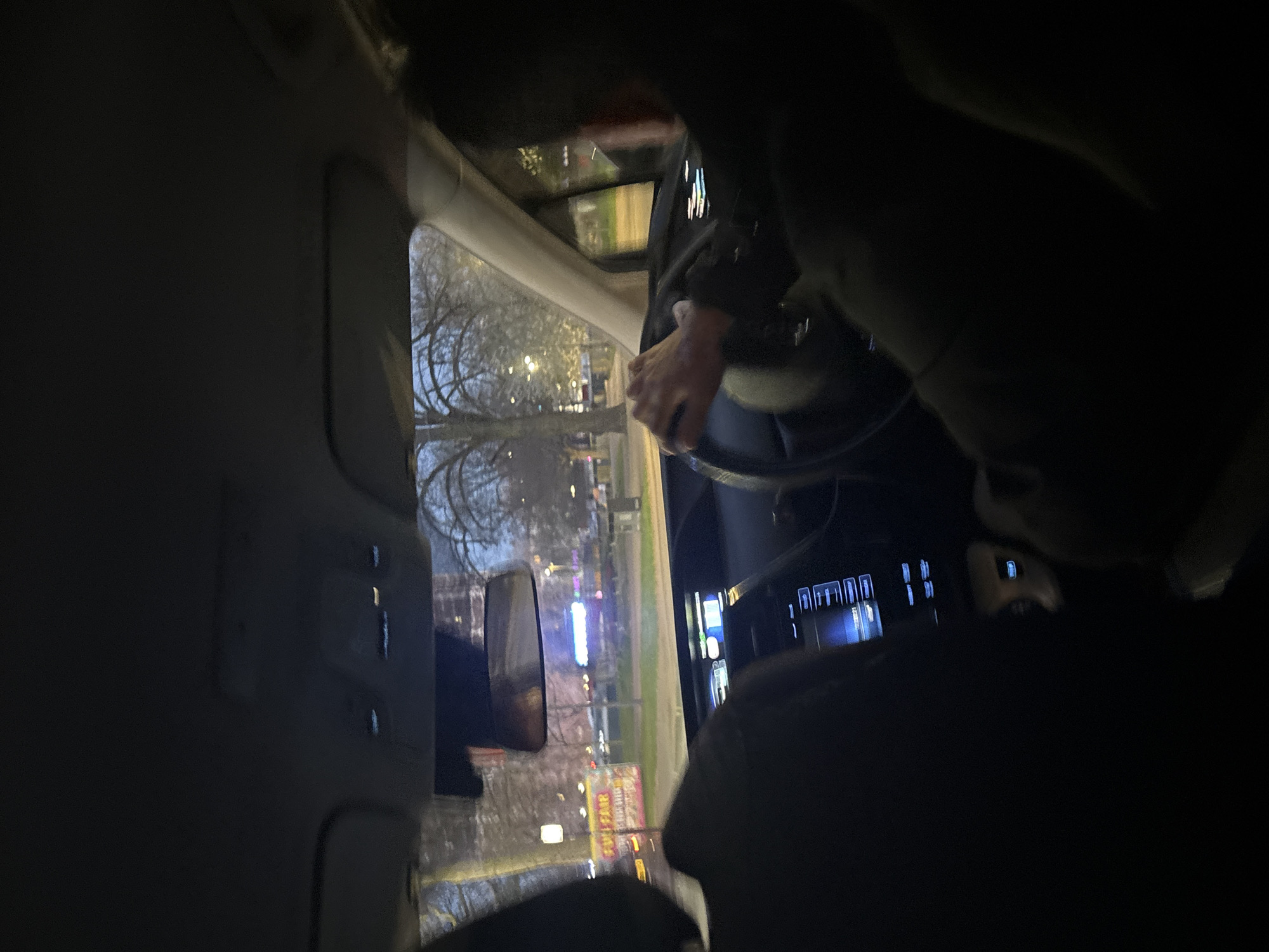 Interior view of a car at night, showing the driver with hands on the steering wheel. Exterior view through the window includes trees, streetlights, and signs, suggesting an urban environment. Visible text on a sign contains "FULL GAME," possibly indicating an arcade or gaming establishment. The location is presumed to be in the city of London, United Kingdom.