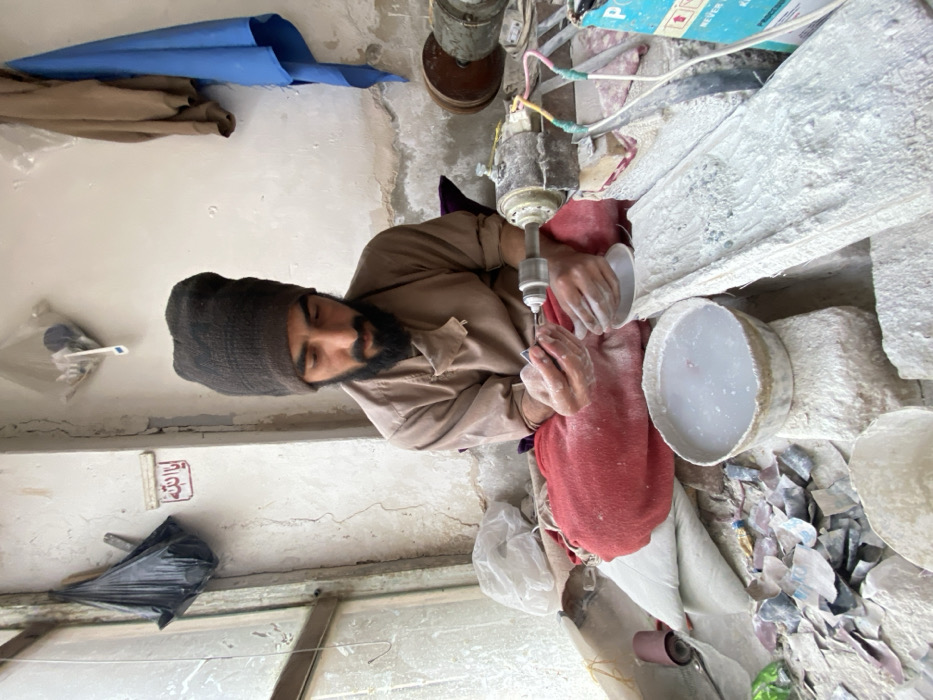 This image depicts an indoor stone crafting workshop in Kandahar, Afghanistan. A man with a dark beard and mustache wears a dark brown knitted cap, a light brown long-sleeved tunic, and red fabric wrapped around his lower body, sitting cross-legged on a dusty floor. He is engaged in working on a small, light-colored stone object held in his left hand, utilizing a rotary tool with a grinding bit operated by his right hand. A bowl filled with murky white liquid, likely water mixed with stone dust, rests directly in front of him. The immediate workspace is covered in light-colored stone dust and scattered fragments of various colored materials, including blue, white, and pink chips. To the man's right, a metal motor assembly with exposed electrical wires sits on a dusty wooden surface, adjacent to a partially visible blue and white box. The background features a grimy window on the left, with a black plastic bag hanging from a hook on the wall below, and a white plaque inscribed with red Arabic script ("Ya Allah"). The wall to the right is discolored and cracked, with a brown and a blue fabric draped over another fixture.