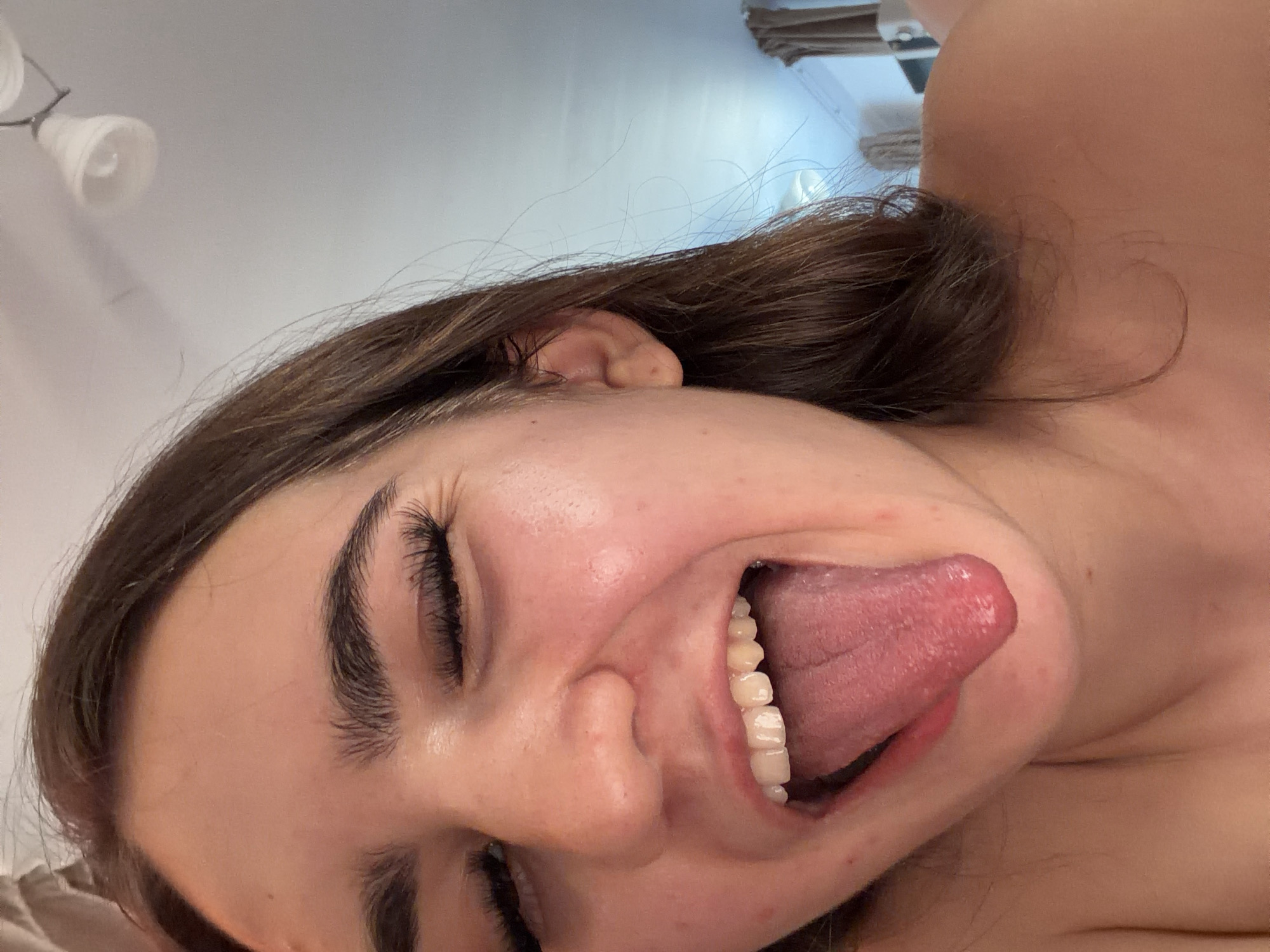 A close-up, tilted shot depicts a young woman with closed eyes, dark hair, and fair skin. Her mouth is open, revealing her tongue and teeth. The background shows a ceiling light and a glimpse of what appears to be a curtain or drape. The image does not contain enough information to establish a location in Voynegovtsi, Bulgaria, or any specific environment.