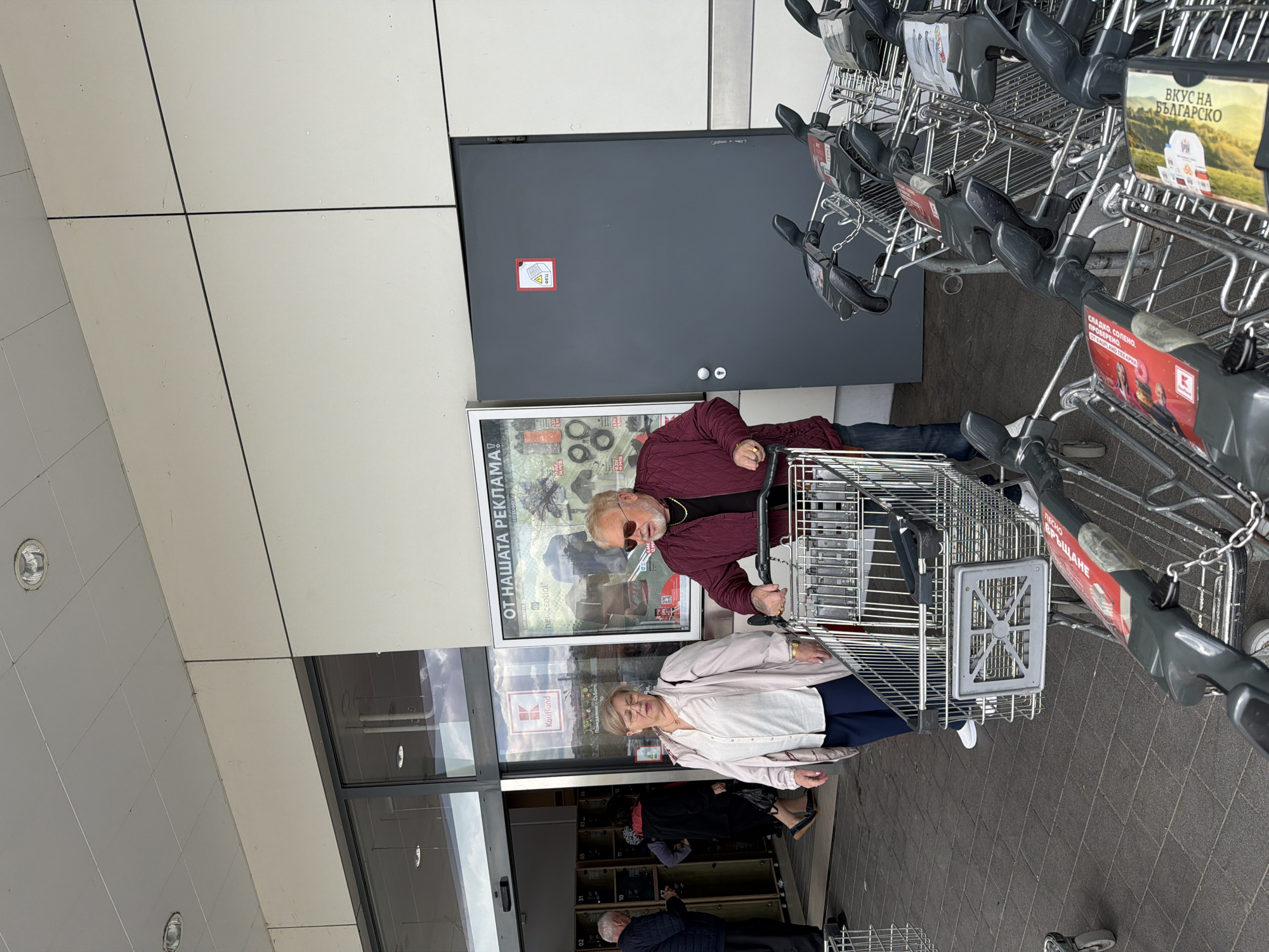 Outside the entrance to a Kaufland store, an elderly couple is standing with a shopping cart filled with items. The man, wearing a maroon jacket and sunglasses, is holding the cart's handle. The woman, wearing a light pink jacket and dark pants, stands beside him. Behind them, other people are entering the store. Several rows of shopping carts are lined up next to the entrance. A large advertisement for Kaufland is displayed on the wall, and the store entrance doors are visible.