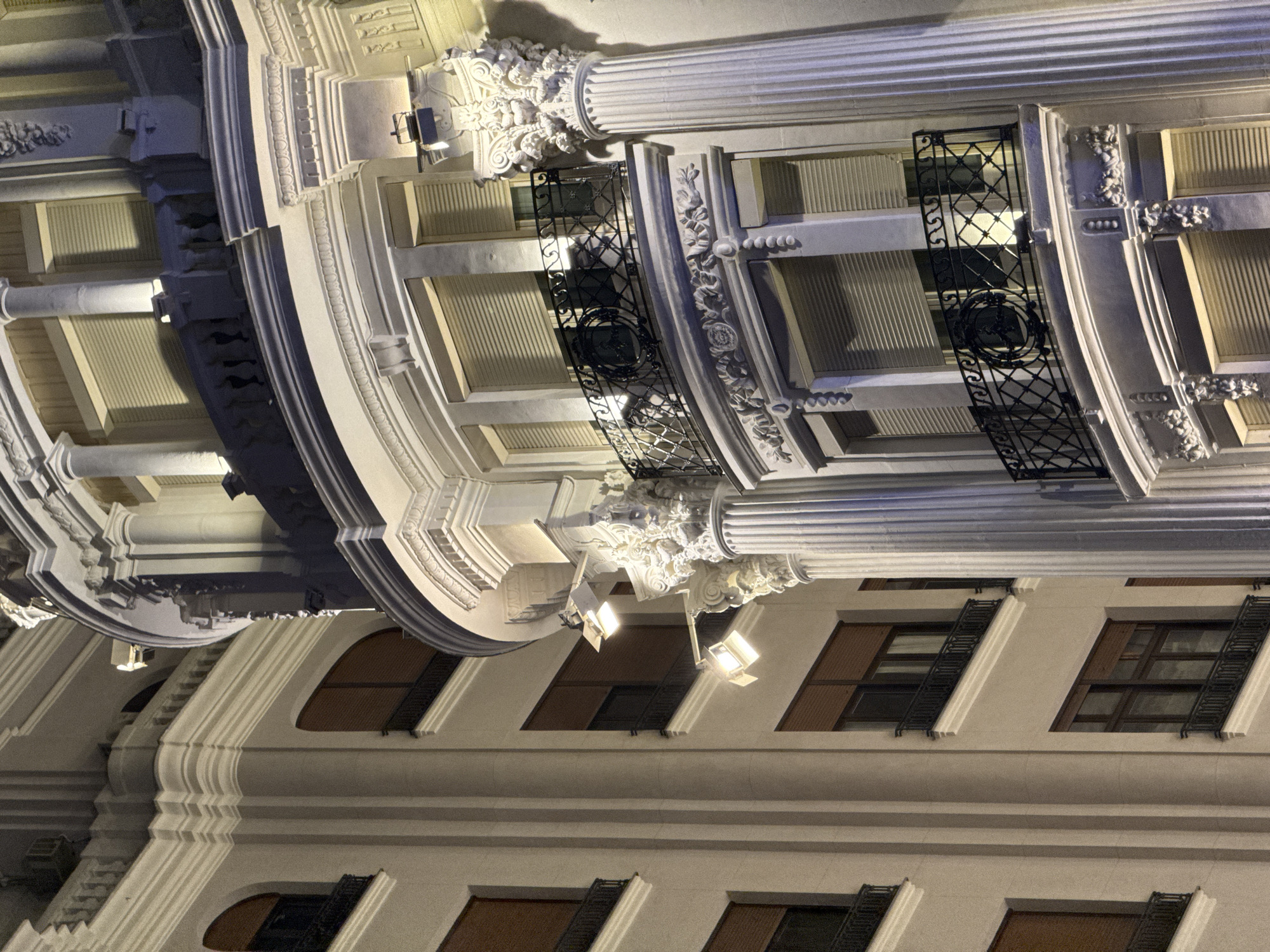 The image shows a nighttime view of the exterior of the Círculo de Bellas Artes building in Madrid, Spain. The architecture is ornate, with classical columns, decorative stonework, and balconies with black iron railings. Multiple windows with closed, slatted blinds are visible. Two floodlights are mounted on the building's facade, illuminating the architectural details. The building appears to be made of light-colored stone or stucco. The lighting creates strong shadows and highlights, emphasizing the three-dimensionality of the structure.