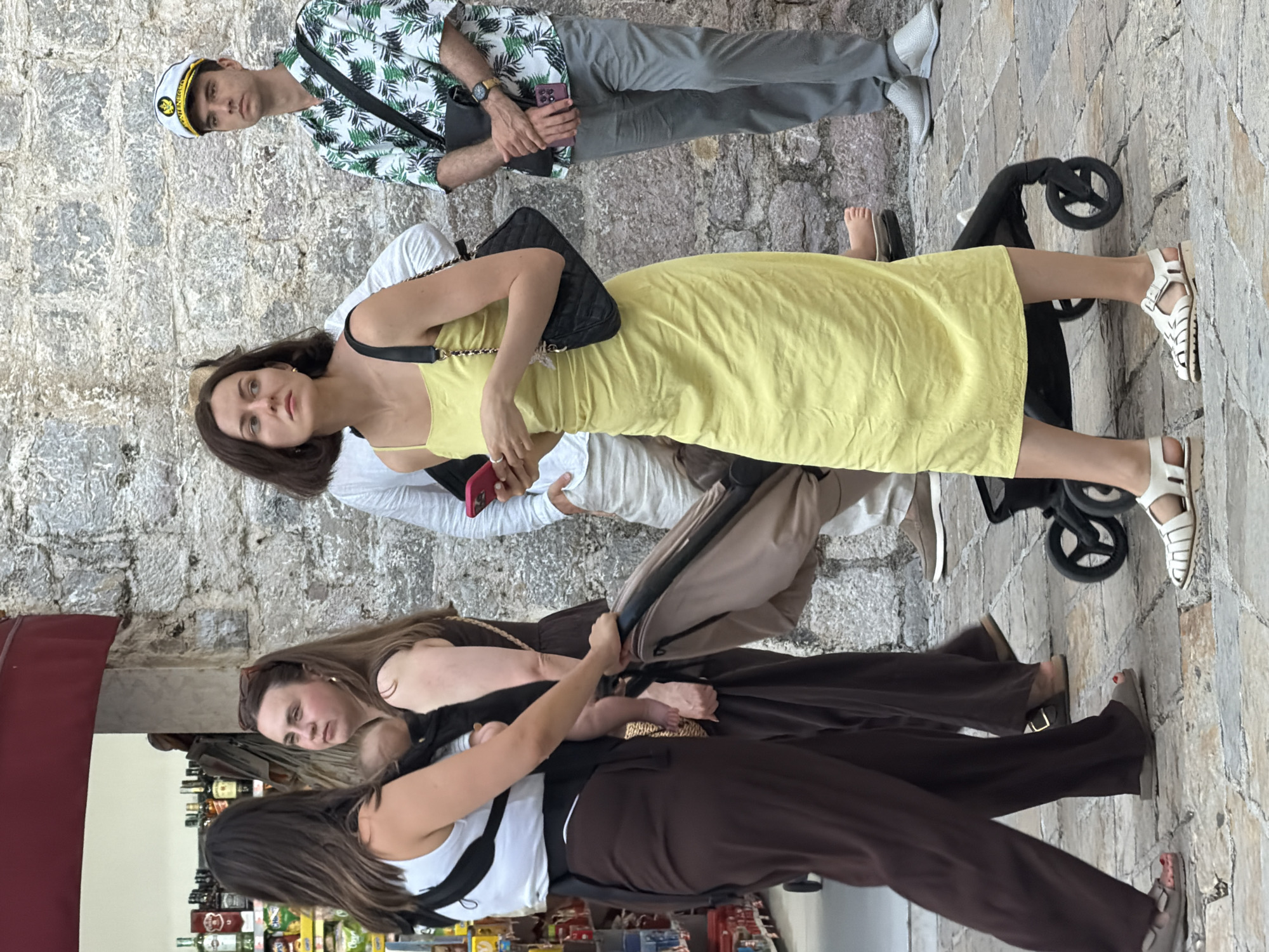 In Budva, Montenegro, a group of people is captured on a cobbled street. To the left, a woman wearing a white tank top and dark brown wide-leg pants carries a baby in a black carrier. Next to her, another woman in a dark dress and sandals pushes a light brown stroller. In the center, a woman in a yellow slip dress holds a pink smartphone and a black quilted purse. Behind her, a man in a white long-sleeved shirt stands partially obscured. To the right, a man in a white captain's hat with "MONTENEGRO" embroidered on it, a green and white tropical print shirt, and gray pants, stands with his arms crossed and a black bag slung over his shoulder. He holds a purple smartphone. The background features a textured stone wall and a glimpse of a storefront with bottles on shelves. The lighting suggests it is daytime.