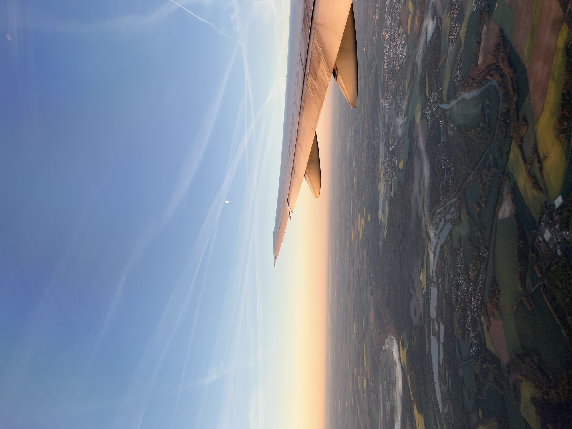 The image shows an aerial view from an airplane, with a wing of the plane visible in the foreground. The sky transitions from blue with contrails to a golden hue near the horizon. Below, a landscape unfolds with a mix of fields, forests, and waterways, likely representing a rural or suburban area. The scene is likely in the vicinity of Congis-sur-Thérouanne, France, based on the landscape characteristics. Details of the city itself are not visible, but fields and a winding river or canal are apparent. There are no people or discernible objects in the immediate landscape visible.