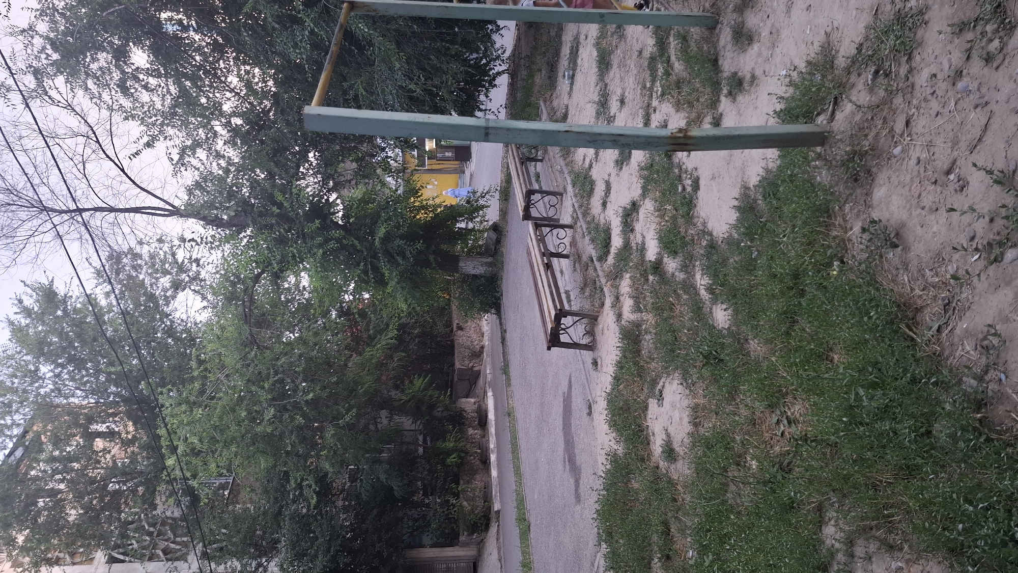 The image depicts an outdoor scene in Shymkent, Kazakhstan, likely in a park or communal area. The primary subjects are two park benches, one positioned in the foreground and another further back. The benches appear to be made of wood for the seating and backs, with decorative metal frames featuring a scrolled design for the legs and supports.
The setting is an urban or suburban environment, with a paved pathway running alongside the benches. To the left of the pathway, dense green foliage and trees dominate the scene, obscuring some of the background buildings, one of which is partially visible and seems to be a multi-story structure. To the right of the pathway, the ground is a mixture of dry, sandy soil and patches of green grass and weeds.
In the background, beyond the benches and pathway, there's a glimpse of a building painted a bright yellow. A person wearing a light blue garment is visible near this yellow building, suggesting the presence of activity or people in the area, though their specific actions are unclear. Overhead, a metallic structure, possibly part of playground equipment or a swing set, is visible. The sky appears overcast or it is dusk, as the lighting is muted and the sky is a pale, diffused color. There is no visible text in the image. The overall impression is one of a somewhat neglected but functional public space, offering a place for rest amidst greenery and urban infrastructure.