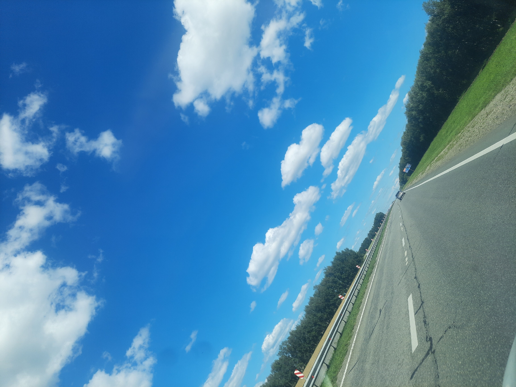 The image depicts a view from a moving vehicle on a highway, looking towards the sky and the side of the road. The sky is a vibrant blue with scattered white clouds. The highway is asphalt with white lane markings. To the left of the road is a metal guardrail with warning signs. Beyond the guardrail, there is a dense line of green trees. The right side of the road features a grassy embankment. In the distance, other vehicles can be seen on the highway. The image is angled, giving a sense of motion and perspective. It is difficult to definitively determine the location in the city of Zvs, Russia, without further context or landmarks.