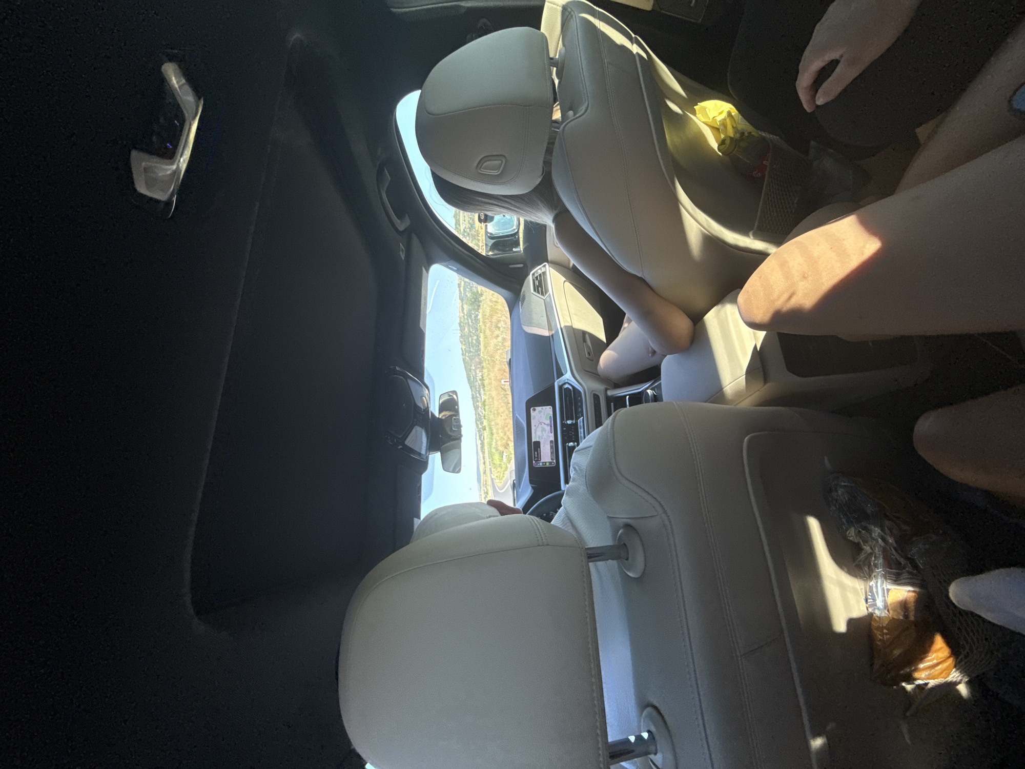 The image shows the interior of a car, looking forward from the back seat. The driver, a person wearing a white shirt, is visible in the driver's seat, operating the vehicle. A passenger, a person with blonde hair, is seated in the front passenger seat, leaning back. The car's interior is furnished with light-colored leather seats. The dashboard features a modern infotainment system displaying a navigation map. Outside the car, a road curves through a landscape of rolling hills and dry vegetation under a clear blue sky. Sunlight streams into the car, creating strong shadows on the seats and floor. In the back seat, the legs of at least two other individuals are visible, one with a prominent sunlit pattern on their leg. A plastic-wrapped item, possibly food, is stored in a mesh pocket on the back of the driver's seat. The scene suggests a road trip on a sunny day. There is no information to suggest the location is Bârlad, Romania.