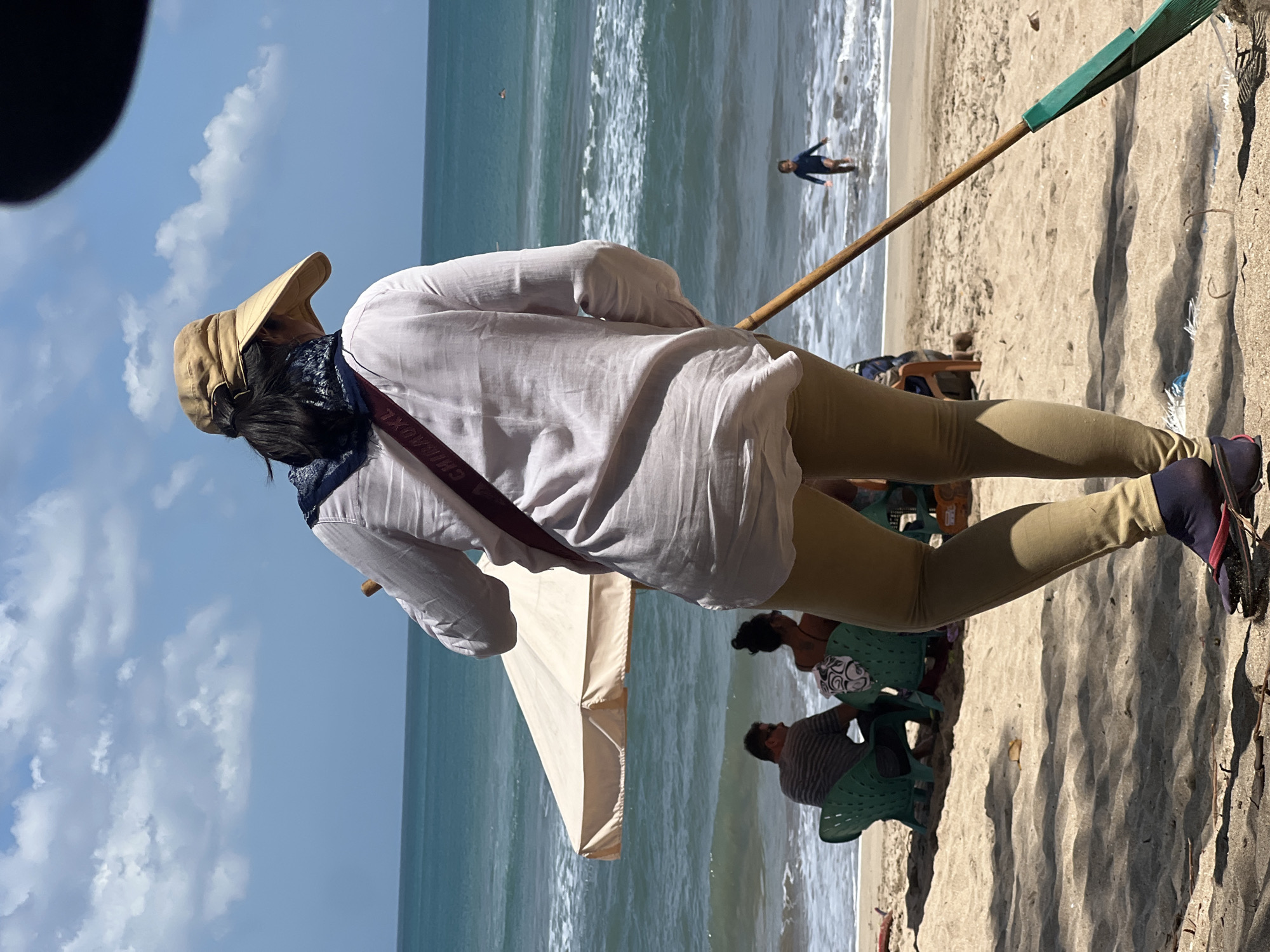 In Kuta, Indonesia, on a sunny beach, a person wearing a light-colored shirt, tan pants, a tan hat, and a bandana around their neck is standing facing the ocean. They are holding a long-handled broom with a green attachment, appearing to sweep the sandy beach. A maroon strap with white lettering crosses their chest. In the background, the turquoise ocean meets the horizon under a blue sky with scattered white clouds. Two individuals are seated on green chairs under a beige umbrella near the water's edge. Further out in the ocean, a small child in a dark blue garment wades in the shallow water. The sand in the foreground is lightly shaded by unseen objects.