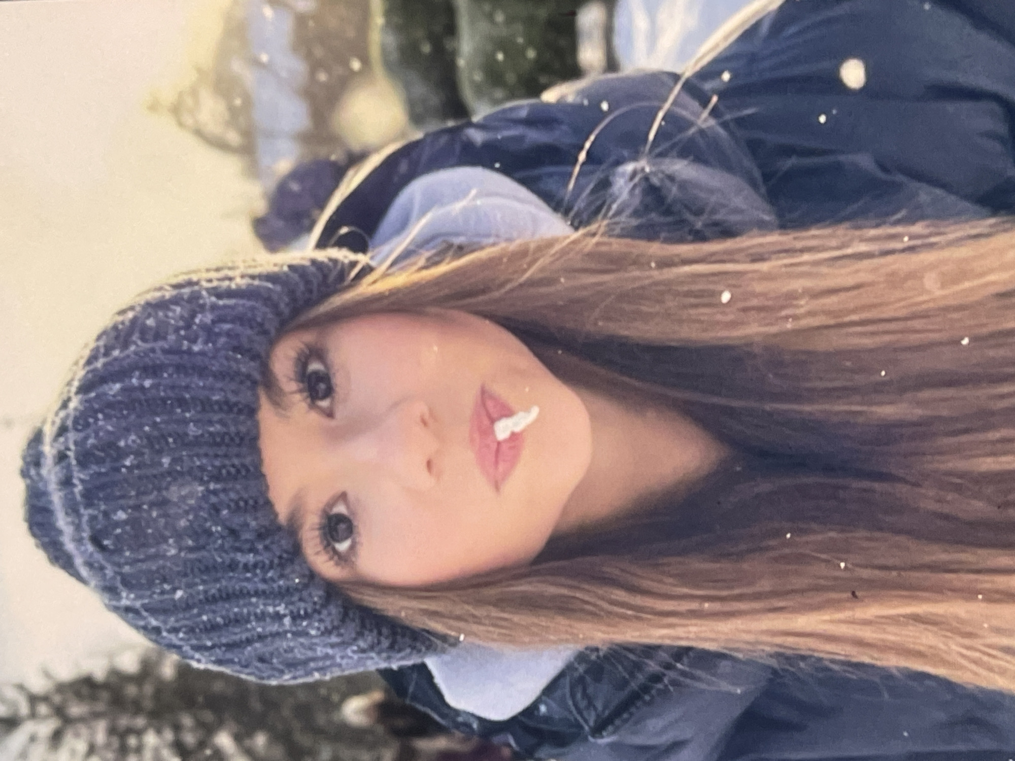 A young woman is captured in a close-up, head-and-shoulders portrait, set outdoors in a snowy winter environment, possibly in Poltava, Ukraine. She looks directly at the camera with a soft, neutral expression. She has long, straight brown hair, with some strands visibly flecked with snowflakes. She is wearing a dark blue or grey knitted beanie hat, also covered in small white snow particles, and a dark blue winter jacket.
A distinctive detail is a small, white, fluffy substance, likely snow or a similar prop, playfully positioned on her upper lip, suggesting she might have just caught or tasted a snowflake. Numerous small white specks are visible throughout the image, indicating falling snow, which has settled on her clothing and hair. The background is softly blurred, showing muted tones of white and darker elements, consistent with snow-covered trees or landscape. The lighting appears soft and even, typical of an overcast winter day. There is no visible text within the image. The scene evokes a quiet, cold, yet somewhat playful moment in a snowy setting.