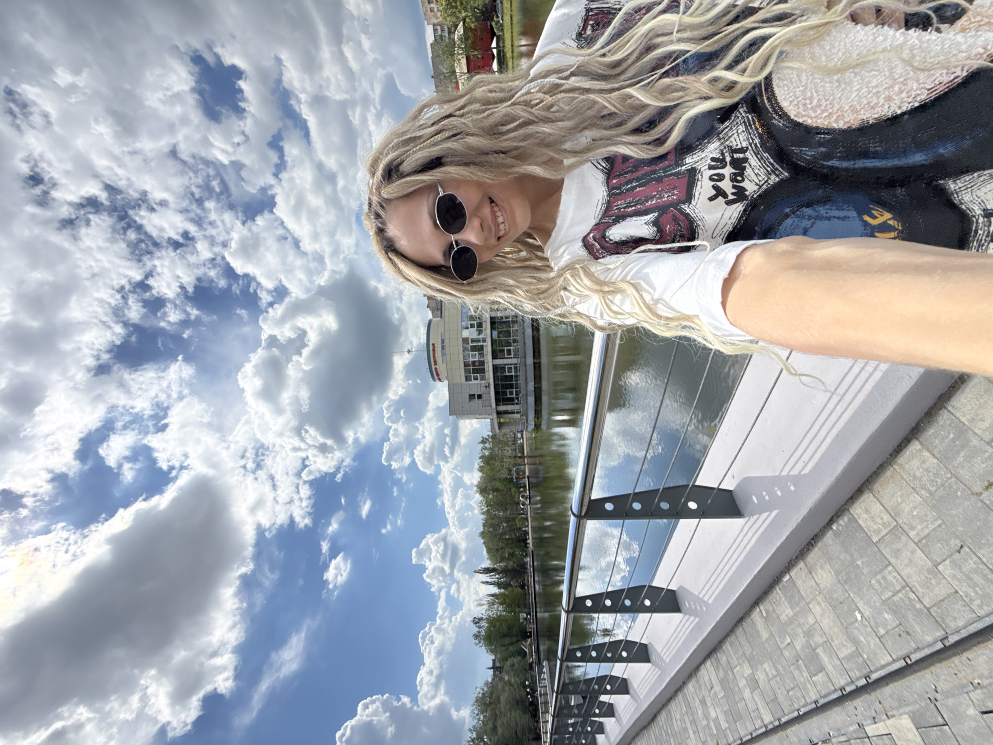 The image is a bright, outdoor selfie featuring a smiling woman on a sunny day in Rusanovo, Russia.
**Subjects:** The primary subject is a young woman, likely taking the photo herself, as evidenced by her extended arm visible in the foreground. She has long, blonde dreadlocks or elaborate braids that cascade around her shoulders. She wears dark, round sunglasses and a white t-shirt with a large, colorful graphic design.
**Scene and Setting:** The woman is positioned on what appears to be a paved promenade or walkway, likely by a river or canal. A contemporary metal railing with multiple horizontal bars separates the walkway from the body of water. Across the water, a line of green trees is visible, partially obscuring a modern-looking, multi-story building with numerous windows.
**Activity:** The woman is actively taking a selfie, smiling broadly at the camera.
**Notable Details:**
* **Time of day/Weather:** The clear blue sky adorned with large, fluffy white clouds indicates it is daytime with bright sunshine. The reflections on the water also confirm a sunny atmosphere.
* **Emotions:** The woman's wide smile suggests happiness or enjoyment.
* **Visible Text:** On her white t-shirt, part of the graphic design includes the white text "YOU WANT".
* **Orientation:** The original image is captured in a portrait orientation but is displayed rotated 90 degrees counter-clockwise.
The setting appears to be a pleasant urban or suburban waterfront area.