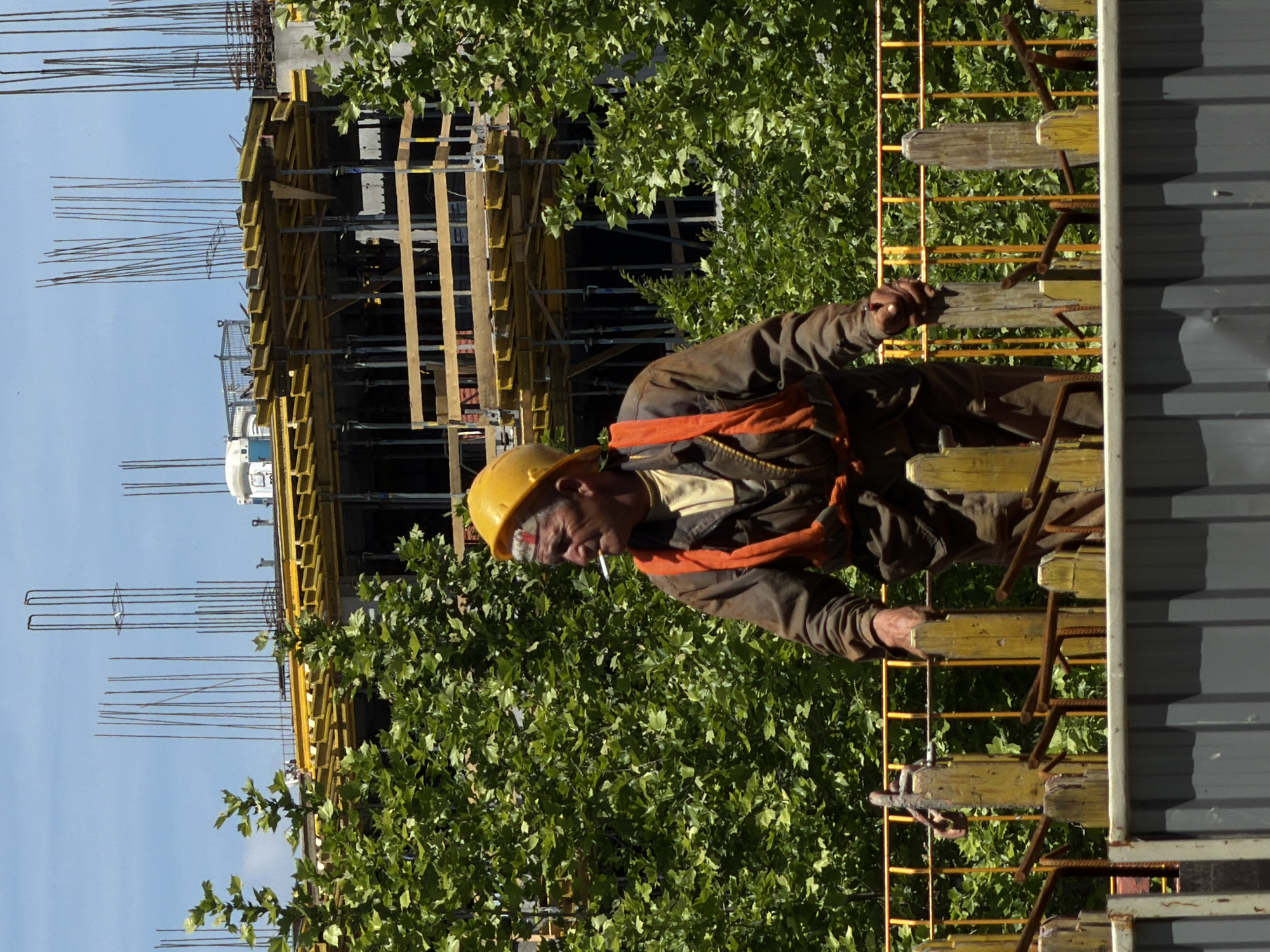 A medium shot of a construction worker in Belgrade, Serbia, smoking a cigarette in his mouth while on a construction site. He wears a yellow hard hat, a brown jacket, and an orange safety harness. He stands on a yellow metal frame with wooden planks, next to a gray metal structure. Lush green foliage surrounds the worker and construction site. The background shows a partially completed building with exposed rebar. The sky is pale blue.