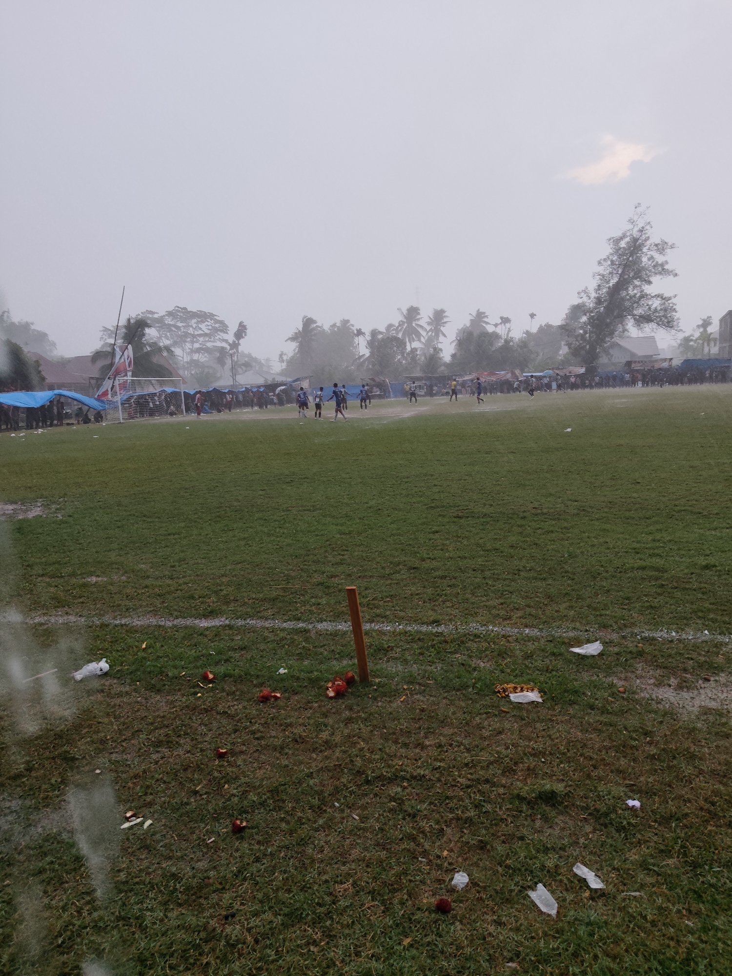The image depicts a soccer field in Meucat, Indonesia, during what appears to be a rain shower. In the foreground, the grass is wet and littered with debris, including red fruits, plastic wrappers, and a discarded white bag. A wooden post stands near the right edge of the frame. In the midground, players in blue uniforms are visible on the field, engaged in a soccer match. Spectators are gathered on the sidelines, sheltered under blue tarps, with a large crowd forming a backdrop. Palm trees and other vegetation frame the scene, with houses and buildings visible in the distance. The sky is overcast and appears hazy due to the rain.