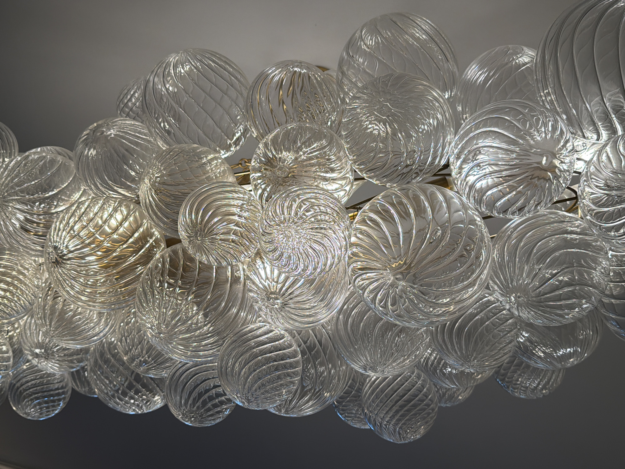 A close-up, low-angle shot captures a cluster of clear, decorative glass orbs, resembling blown glass art or components of a chandelier. The orbs vary in size and are arranged densely, creating a complex, textured surface. Each orb features intricate swirling patterns molded into the glass, which catch and refract the light, producing highlights and shadows that give them a dynamic, almost liquid appearance. Some orbs are partially illuminated from within, suggesting internal light sources or the reflection of ambient light. The orbs are suspended from thin, metallic rods, likely part of a light fixture. The background is a soft, neutral gray, allowing the orbs to be the sole focus of the image. The overall impression is one of elegance and sophisticated design, with the interplay of light and textured glass creating a visually captivating effect. This image does not depict a scene in San Marino, United States of America.