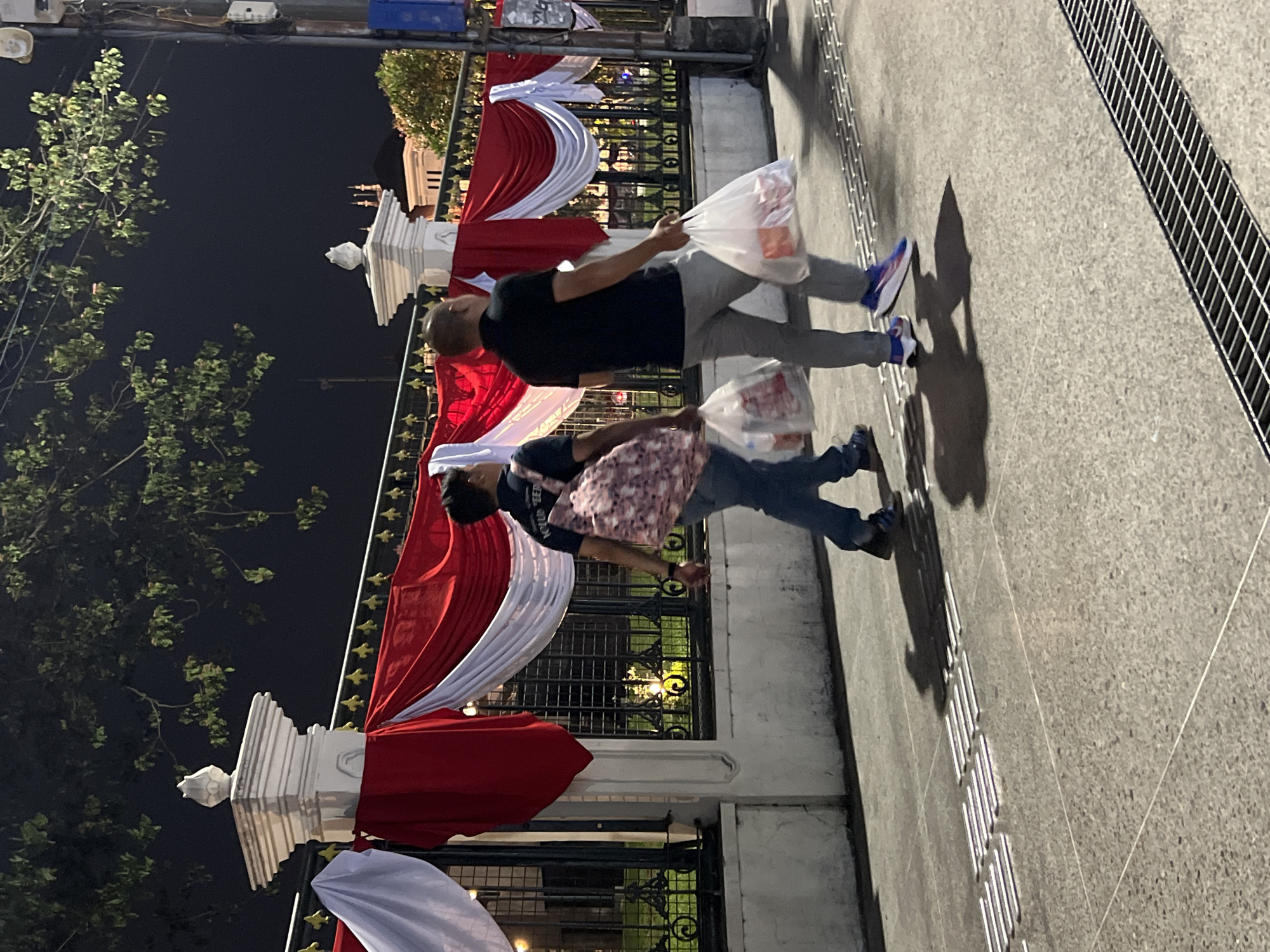 Two individuals walk on a paved sidewalk at night, with red and white draped fabric decorating a fence in the background. The first individual, on the left, wears a dark blue t-shirt and jeans, carrying a patterned backpack and a white plastic bag. The second individual, on the right, is dressed in a black polo shirt and grey trousers, also carrying a white plastic bag. Both are walking away from the camera. The environment suggests an outdoor urban setting, possibly a public space in Yogyakarta, Indonesia, given the prominent use of red and white drapery, which are the colors of the Indonesian flag and frequently used for national celebrations or events in Indonesia. A street light is visible on the right side of the image.