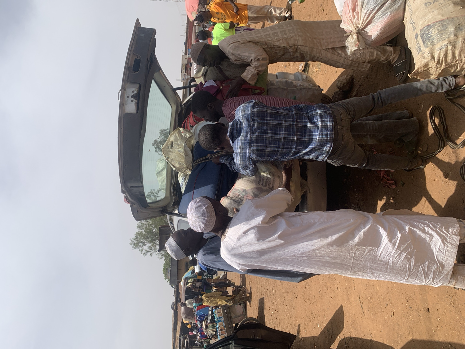 This image captures a bustling outdoor scene in Rafin Par, Nigeria, under bright, clear daylight. The main subjects are a group of approximately ten men, some wearing traditional long robes and skullcaps, while others are dressed in more casual attire like plaid shirts and jeans.
The central activity revolves around a dark-colored car with its trunk open. Several men are actively engaged in loading various items into the car, including large white sacks, which appear to contain goods, and blue luggage bags. Men are seen bending over, reaching into the trunk, and pushing items, indicating a coordinated effort to pack the vehicle. One man in a plaid shirt and jeans is prominently leaning into the trunk, while others assist or observe from around the car.
The setting is characterized by a reddish-brown, dusty ground. Strong shadows cast by the individuals and objects suggest the time of day is likely midday or early afternoon. In the background, beyond the immediate group, other people, structures, and what appears to be market stalls or tents are visible, hinting at a busy public space, possibly a market or a transportation hub. A large white sack in the upper right corner features the visible text "JUGRA". The overall impression is one of purposeful activity and cooperation in a communal environment.