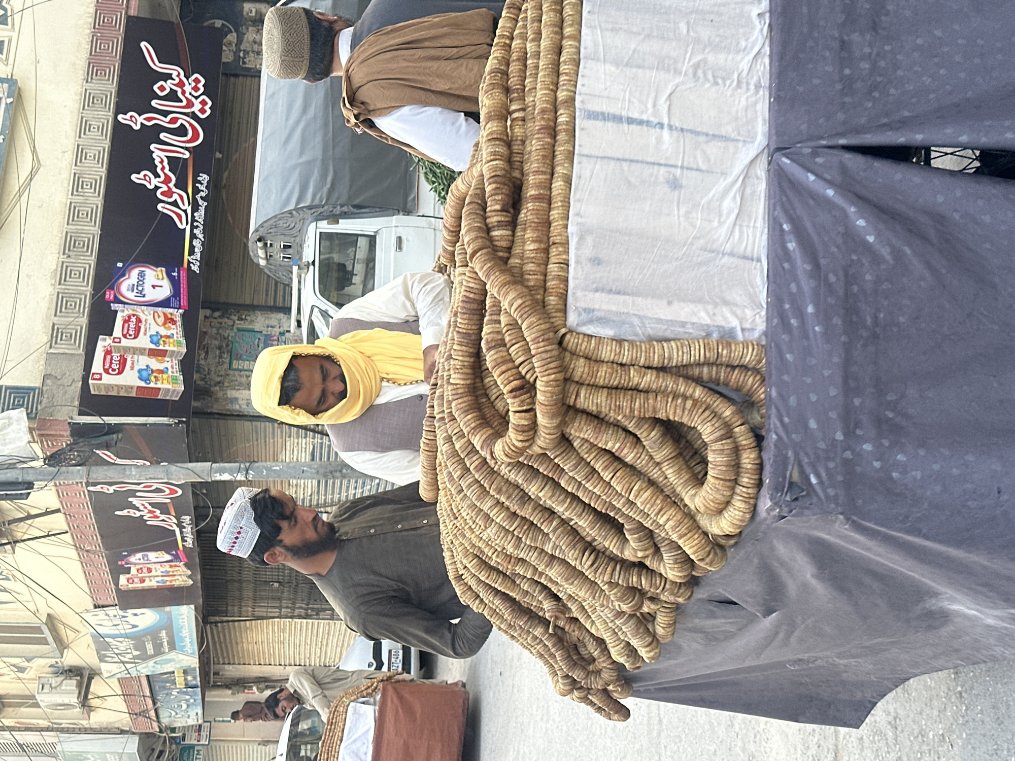 A street scene in Quetta City Tehsil, Pakistan. A man wearing a white and red cap and grey shirt stands next to a cart covered in white and black cloth. The cart is piled with long strings of dried figs. Two other men are standing next to the cart. The first is wearing a yellow head scarf. A storefront is visible in the background with infant formula products on display.