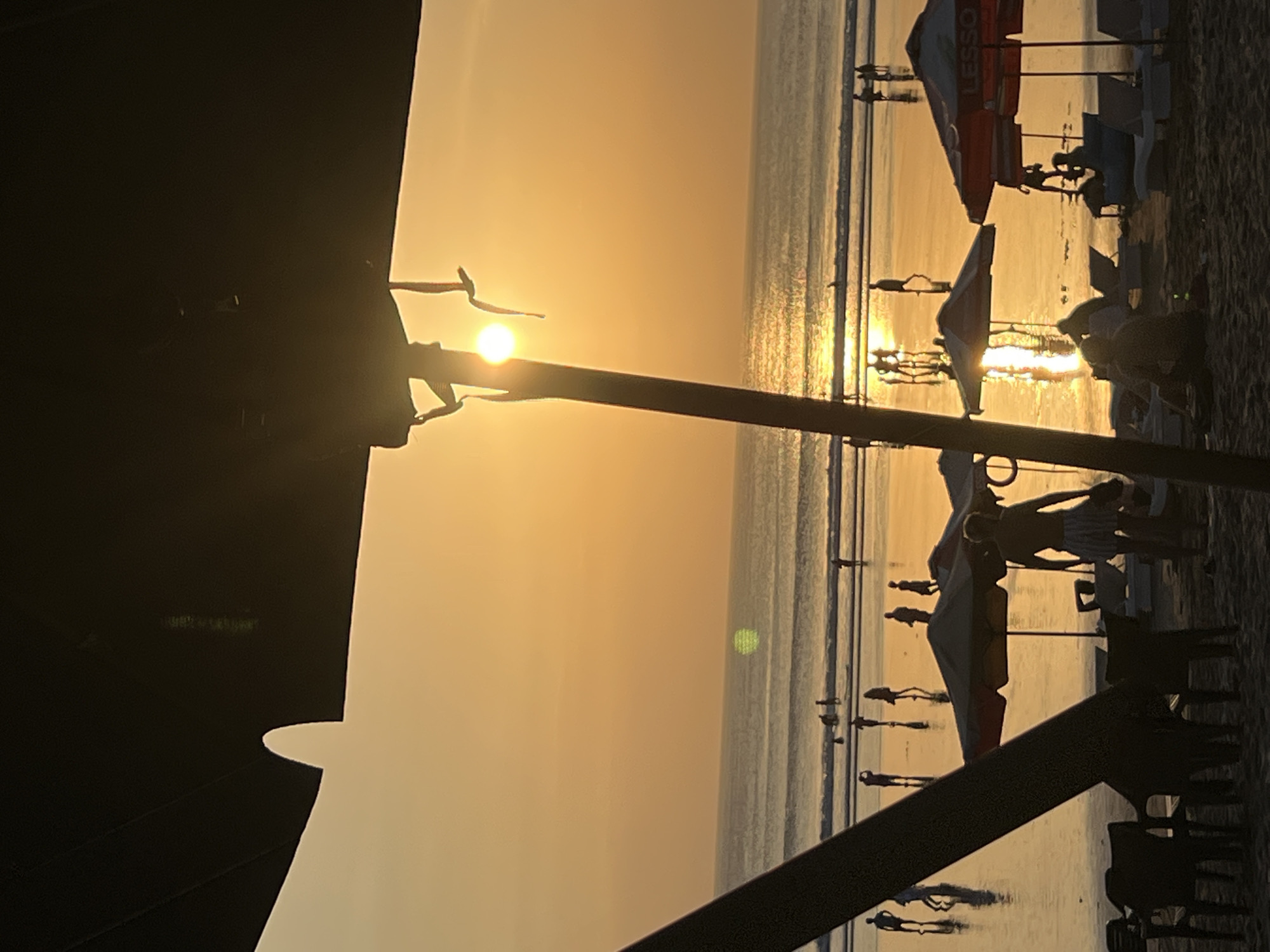 This is a silhouette photograph of Kuta Beach in Bali, Indonesia at sunset. The sun is low on the horizon, casting a warm orange glow over the scene. The ocean stretches out to the horizon, with gentle waves lapping at the shore. Silhouettes of people are visible on the beach, some walking, some standing, and some sitting under umbrellas. The foreground is dominated by the dark silhouette of an umbrella and its pole. The sand is visible in the very bottom of the frame, with chairs and lounge chairs scattered on it. The overall atmosphere is peaceful and serene.