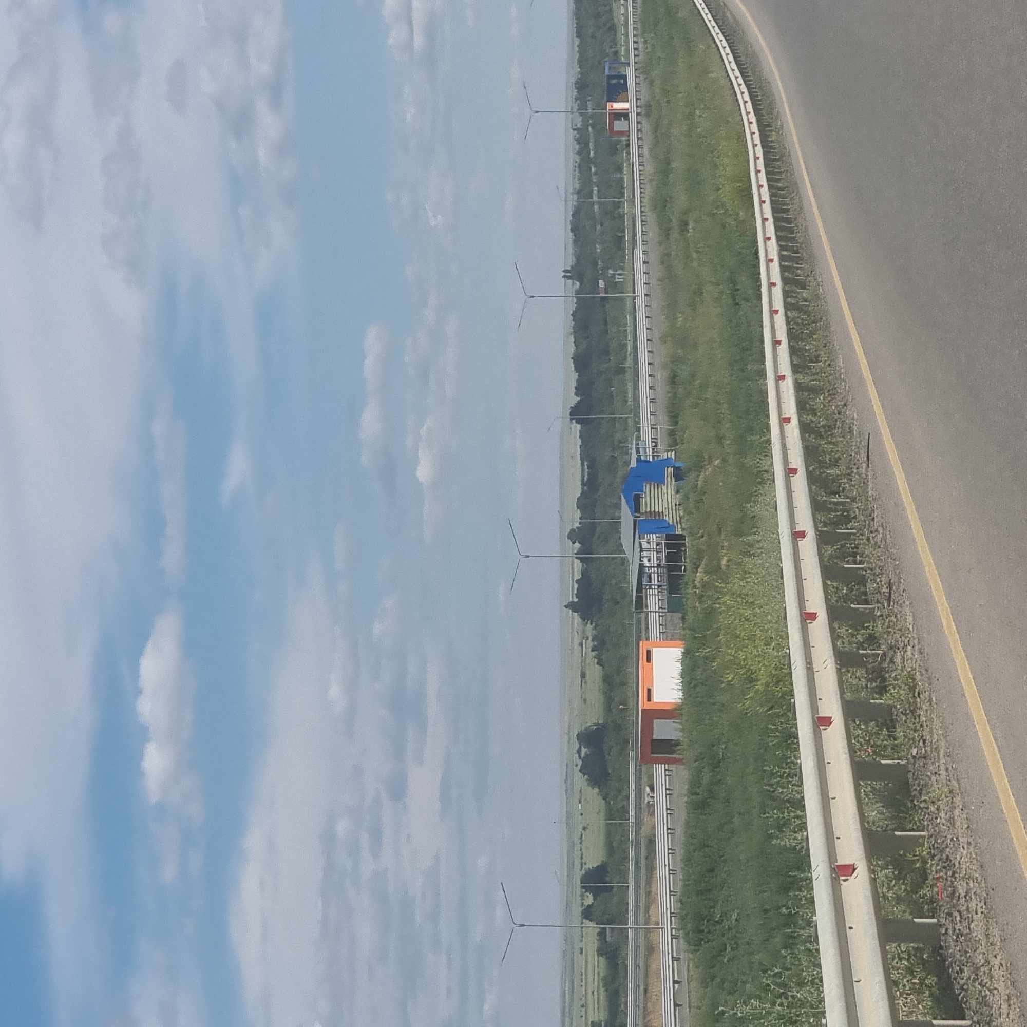 The image shows a highway scene under a cloudy blue sky, likely taken from a moving vehicle. In the foreground, the edge of a paved road is visible with a yellow lane marker. A metallic guardrail with red reflectors runs along the side of the road. Beyond the guardrail, a strip of green grass and vegetation separates the road from the main area.
In the middle ground, there are several structures and infrastructure elements. A long, white guardrail is present on the opposite side of the highway. Two small, orange and white modular buildings are situated near the highway. One appears to be a simple rectangular structure with a white door or panel. Another structure, partially covered with blue tarpaulin and green material, is next to it, resembling an open-sided shelter or construction area with some visible posts and beams. Several streetlights with a modern design are spaced along the highway.
The background depicts a vast, flat landscape with green fields and scattered trees, suggesting a rural or semi-rural environment. The horizon is low, and the sky is filled with scattered white clouds against a blue backdrop. The overall impression is one of an open, developed highway in a predominantly natural, undeveloped setting. The location is identified as Alğa, Kazakhstan.