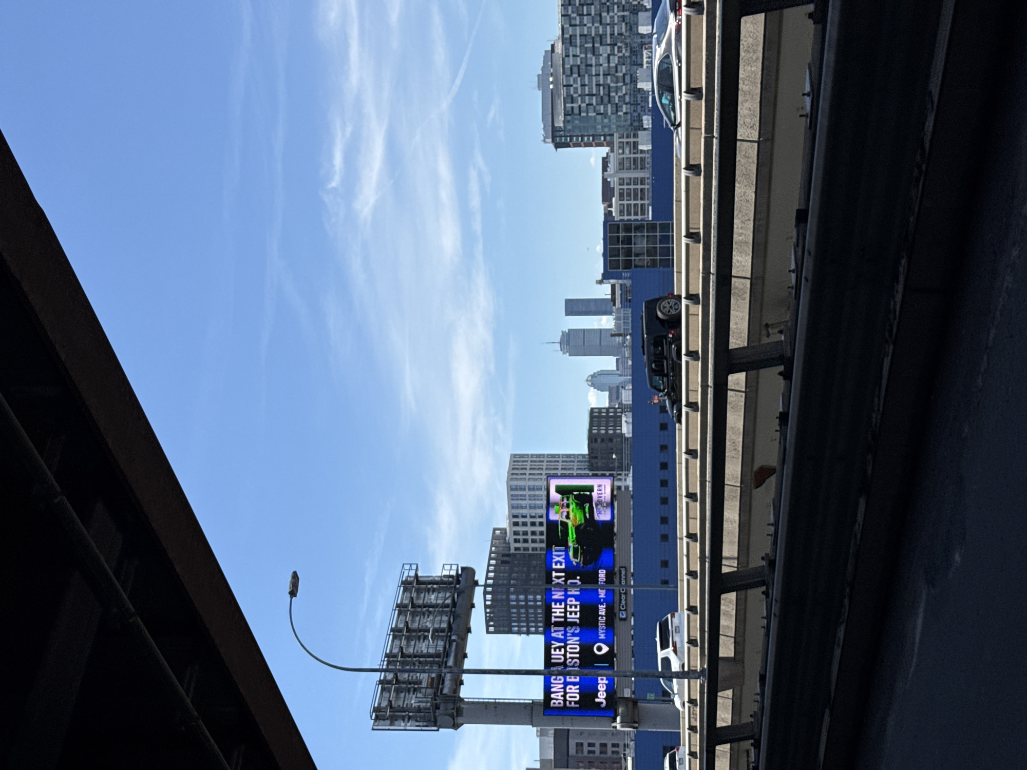 The image depicts an urban scene in Somerville, Massachusetts, USA. A highway overpass dominates the right side, with cars visible driving on it. A view of the city is visible, including buildings and skyscrapers. A large billboard is prominently displayed on the left side of the image, advertising Jeep and Ford. The sky is blue with wispy clouds. A street light and utility poles are also present in the view.