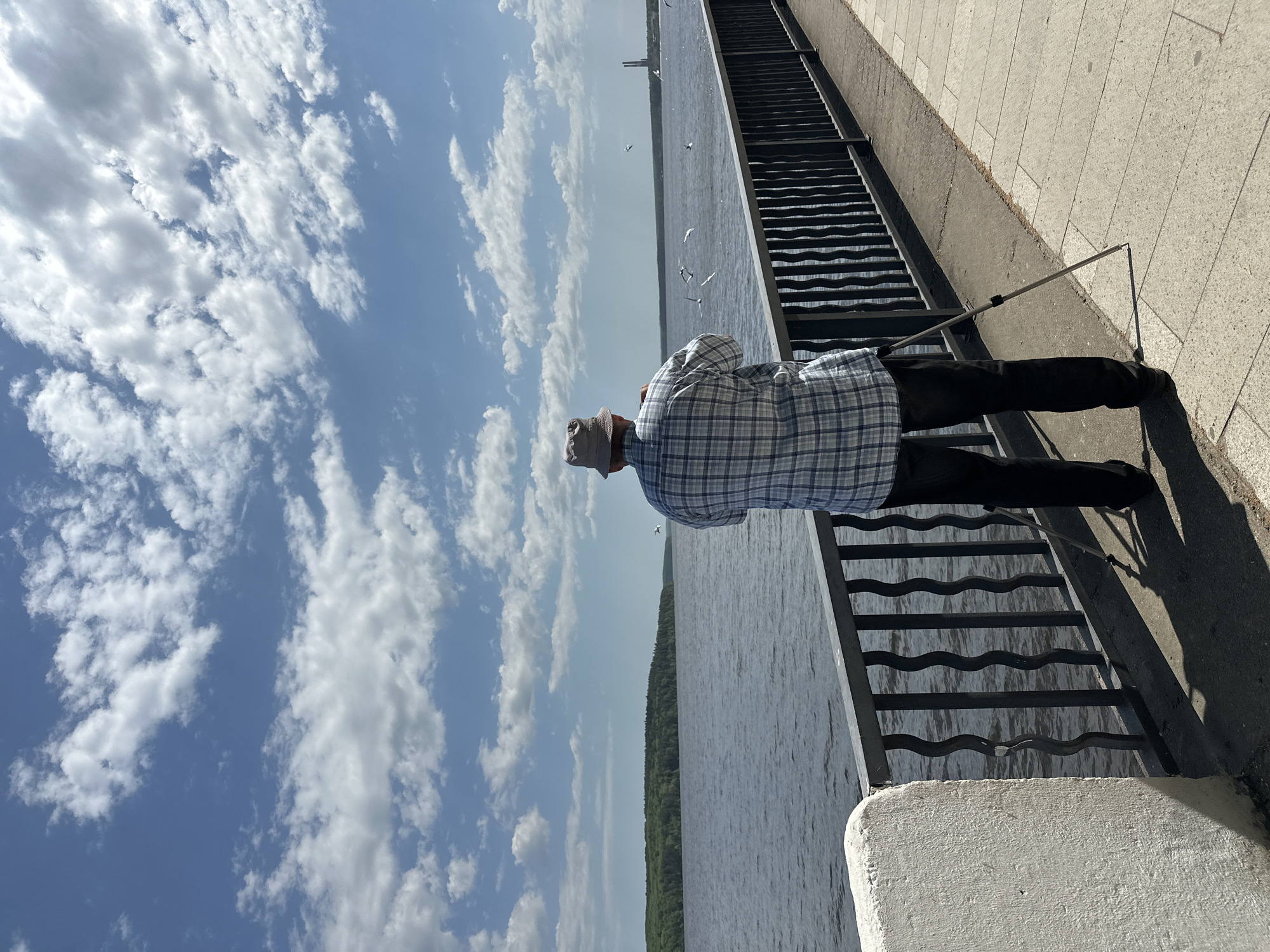 A man stands on a bridge in Chaikovsky, Russia, overlooking a wide body of water. He wears a plaid shirt, dark pants, and a light-colored hat. His arms are crossed, and he appears to be looking out at the water and the sky filled with white clouds. A metal railing runs along the edge of the bridge. A set of stairs leading down to the water is visible.