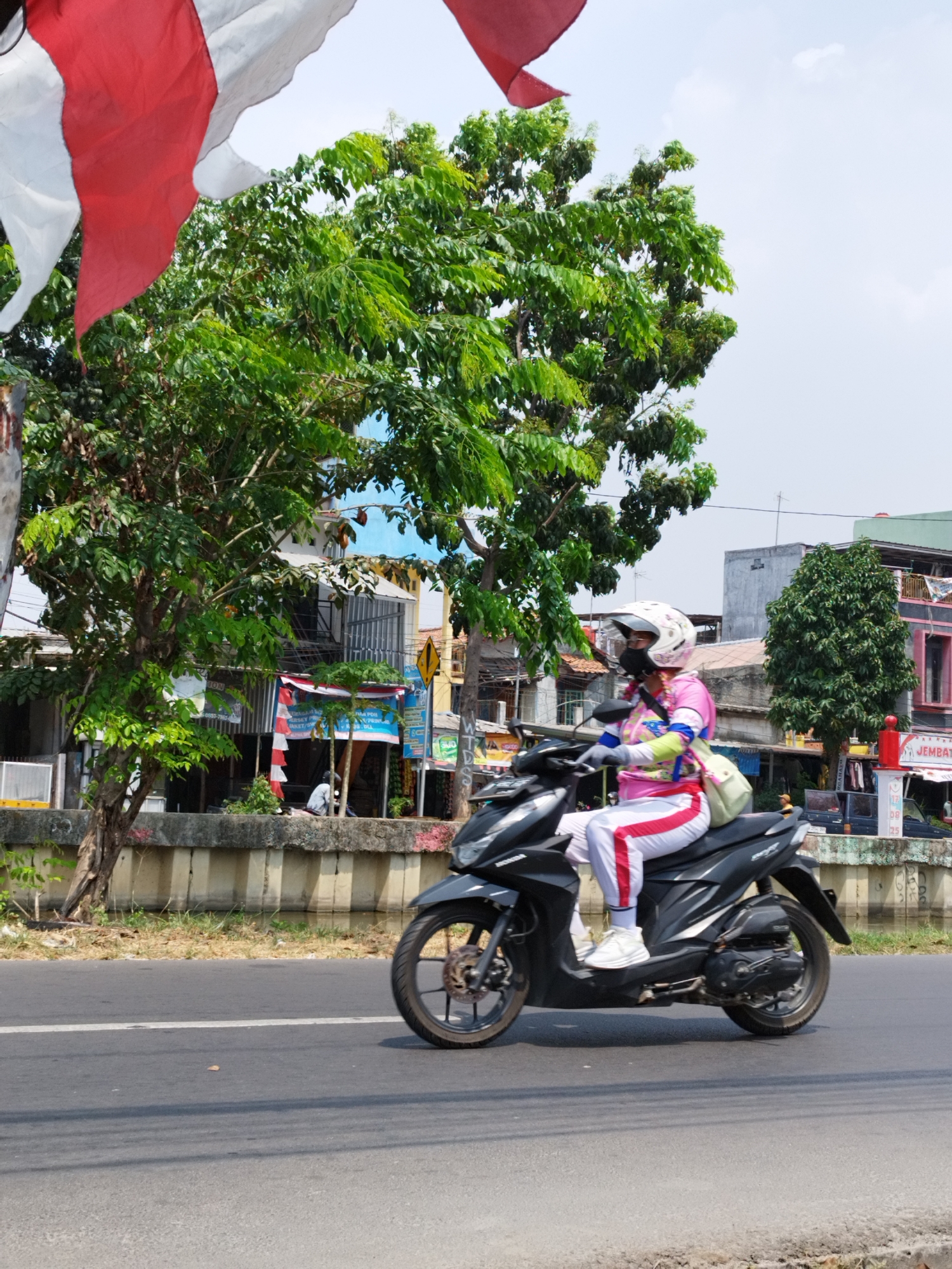 A woman wearing a white helmet, a pink long-sleeved shirt with a colorful design, white pants with red stripes, and white sneakers is riding a black scooter. She is also wearing a light green bag over her shoulder. The scooter is traveling on a paved road with a white line marking its lane. In the background, there are trees, buildings, and a canal. The flags of Indonesia (red and white) are visible at the top left of the image. The scene appears to be in the city of Bekasi, Indonesia. The weather is clear and sunny.