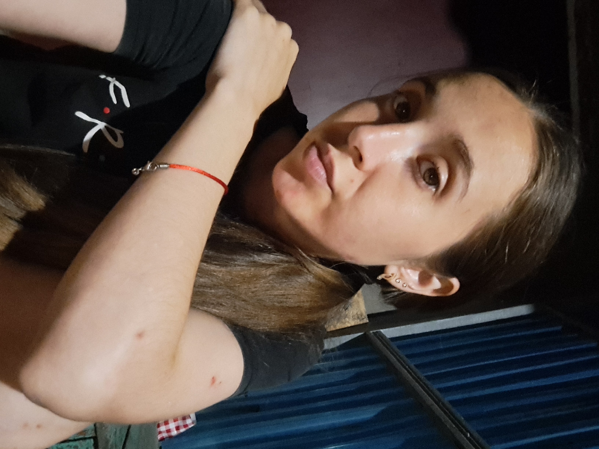 A young woman with long, brown hair, wearing a black t-shirt and a red bracelet, is shown in a close-up, selfie-style shot. She has multiple earrings in her left ear. Her right arm is resting on her left shoulder, and her left arm is bent at the elbow. The background is dark and appears to be an outdoor or semi-outdoor setting, with what looks like corrugated metal siding on the right. There are some minor red marks on her right arm and shoulder. The image is likely a personal photograph taken by the subject herself.