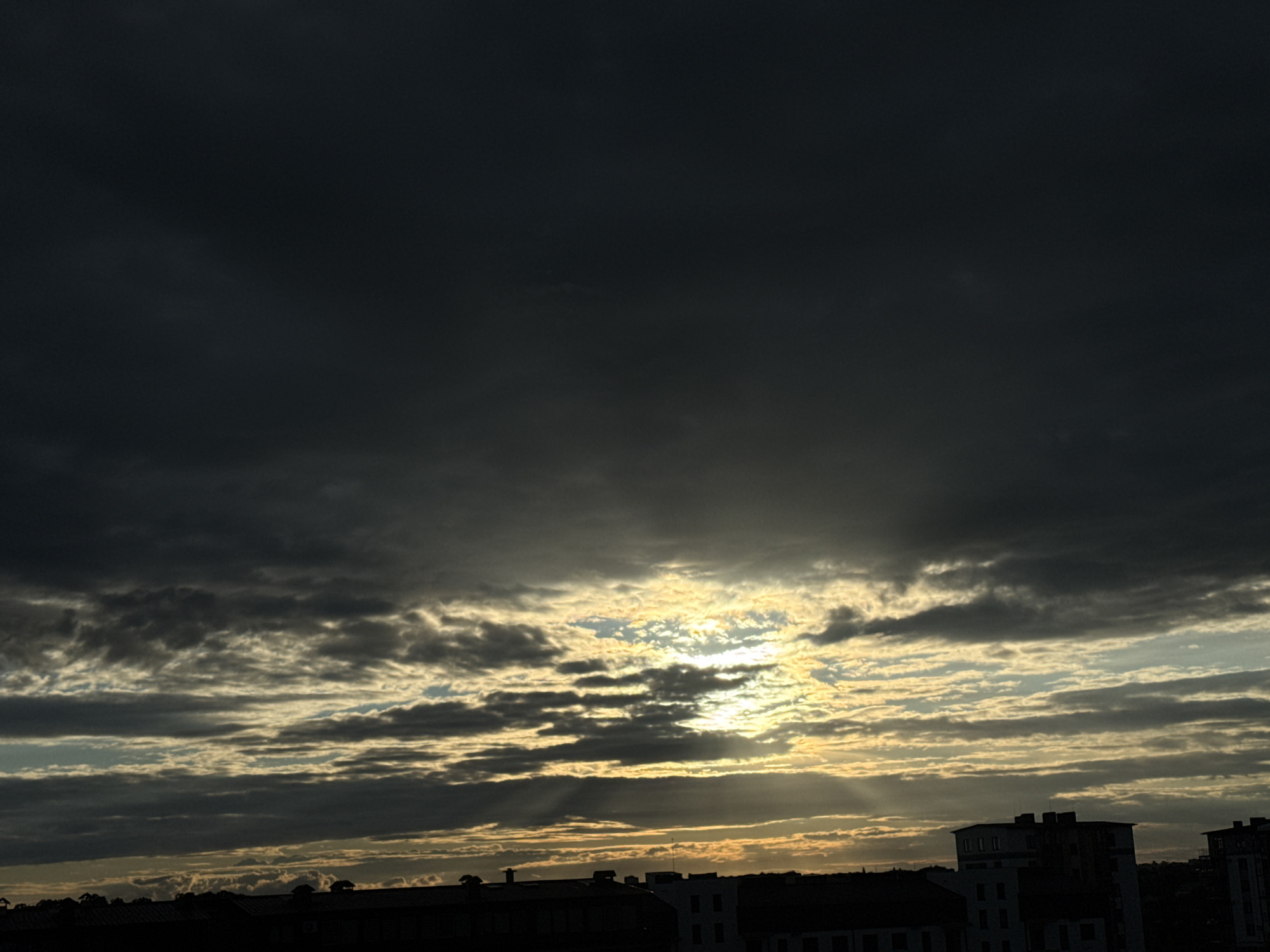 The image depicts a dramatic sky filled with dark, heavy clouds that are partially illuminated by the setting sun. Streaks of sunlight break through the clouds, creating a dramatic effect as they cast light and shadow across the sky. In the foreground, the silhouettes of buildings are visible against the bright sky. These buildings appear to be part of a cityscape, with various rooflines and structures suggesting an urban environment. The overall mood of the image is somber and awe-inspiring, likely captured during twilight or a storm. The location is described as Unknown, Unknown.