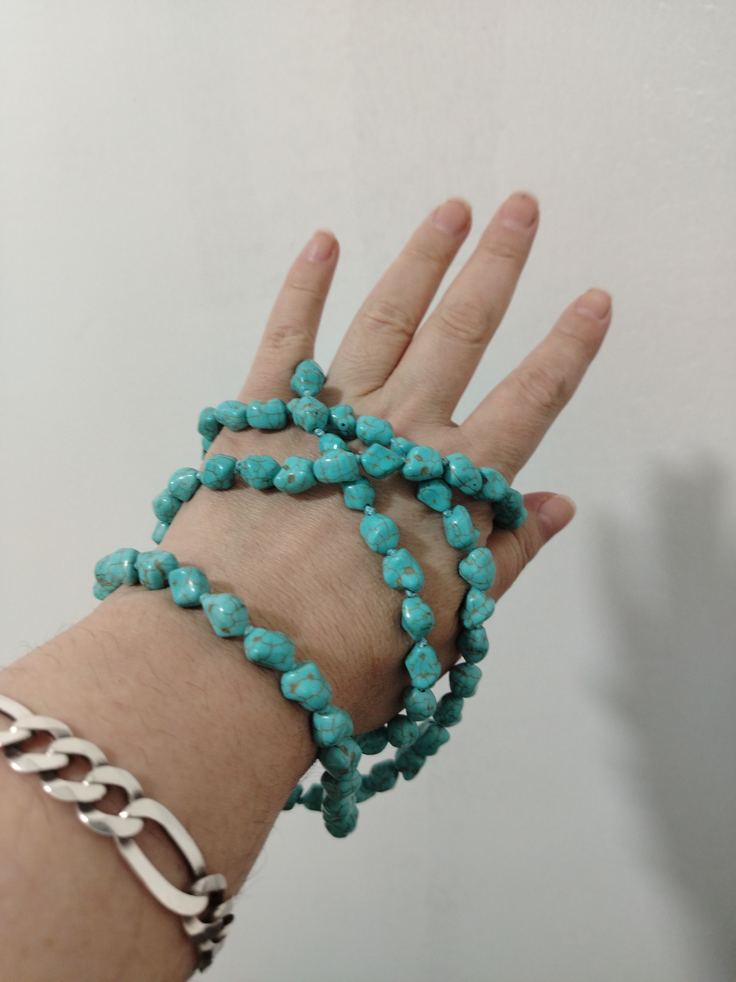 A close-up shot shows a human hand draped with a string of turquoise beads. The hand, seemingly fair-skinned, is adorned with a silver bracelet visible on the wrist. The hand rests against a plain white wall. The location is unknown; the claim that the image was taken in Al Mas'oudi, United Arab Emirates, cannot be verified.
