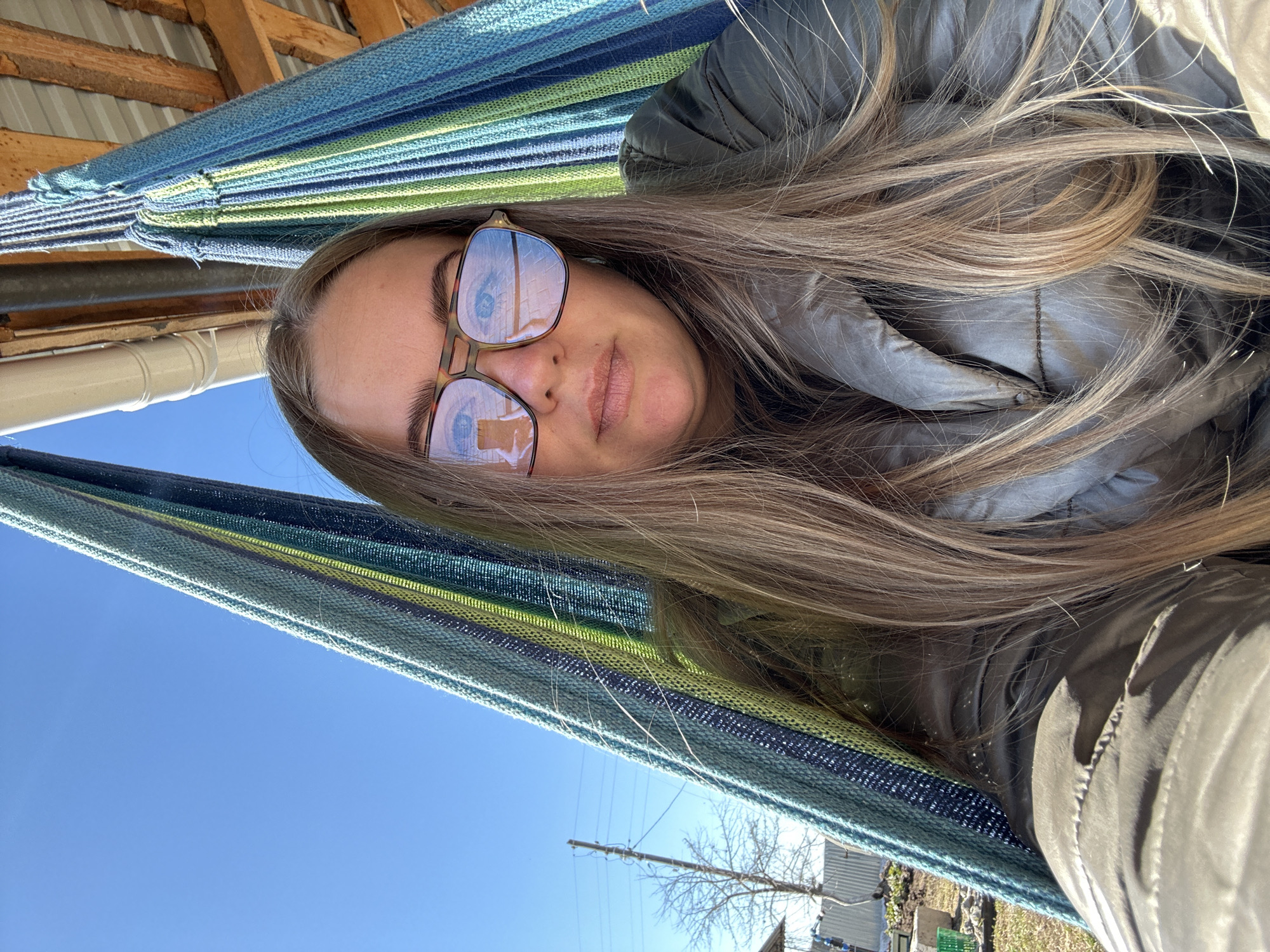 A woman is lying in a striped hammock with a clear blue sky visible in the background. She is wearing sunglasses with a building reflected in the lenses. She has long, light brown hair and is wearing a gray jacket. The hammock is located in Уральское, Russia.