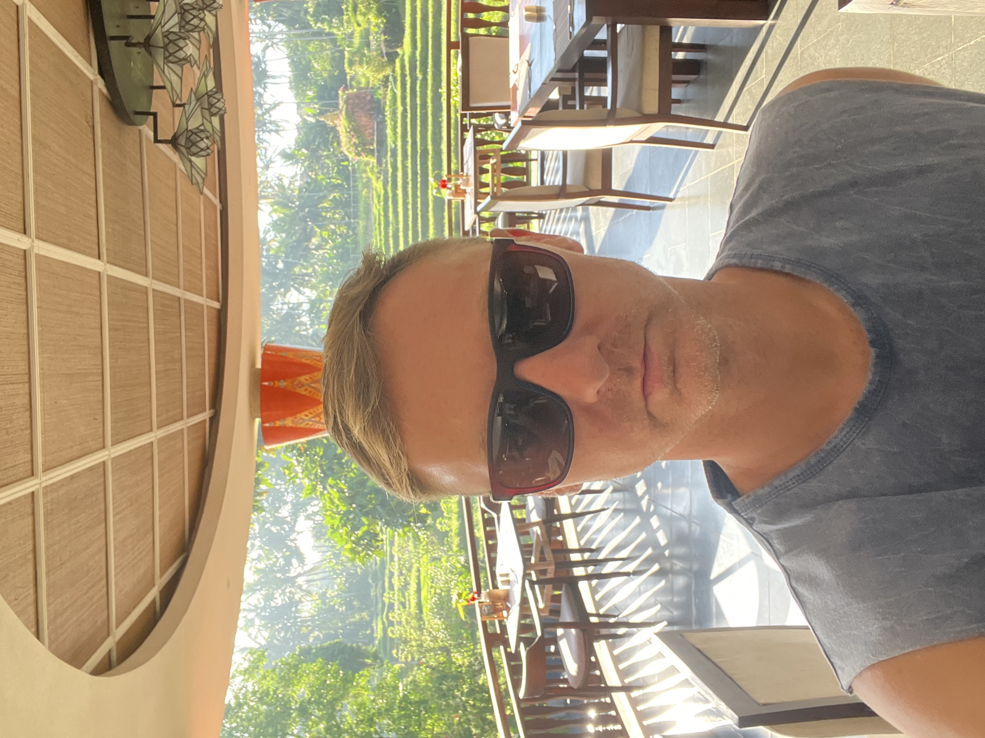 A man wearing sunglasses and a gray t-shirt is taking a selfie in a restaurant overlooking rice terraces. The restaurant is likely located in the Ubud District, Indonesia. Tables and chairs are visible in the foreground. The rice terraces are in the background. A building with a unique, rounded design is visible on the left side of the frame.