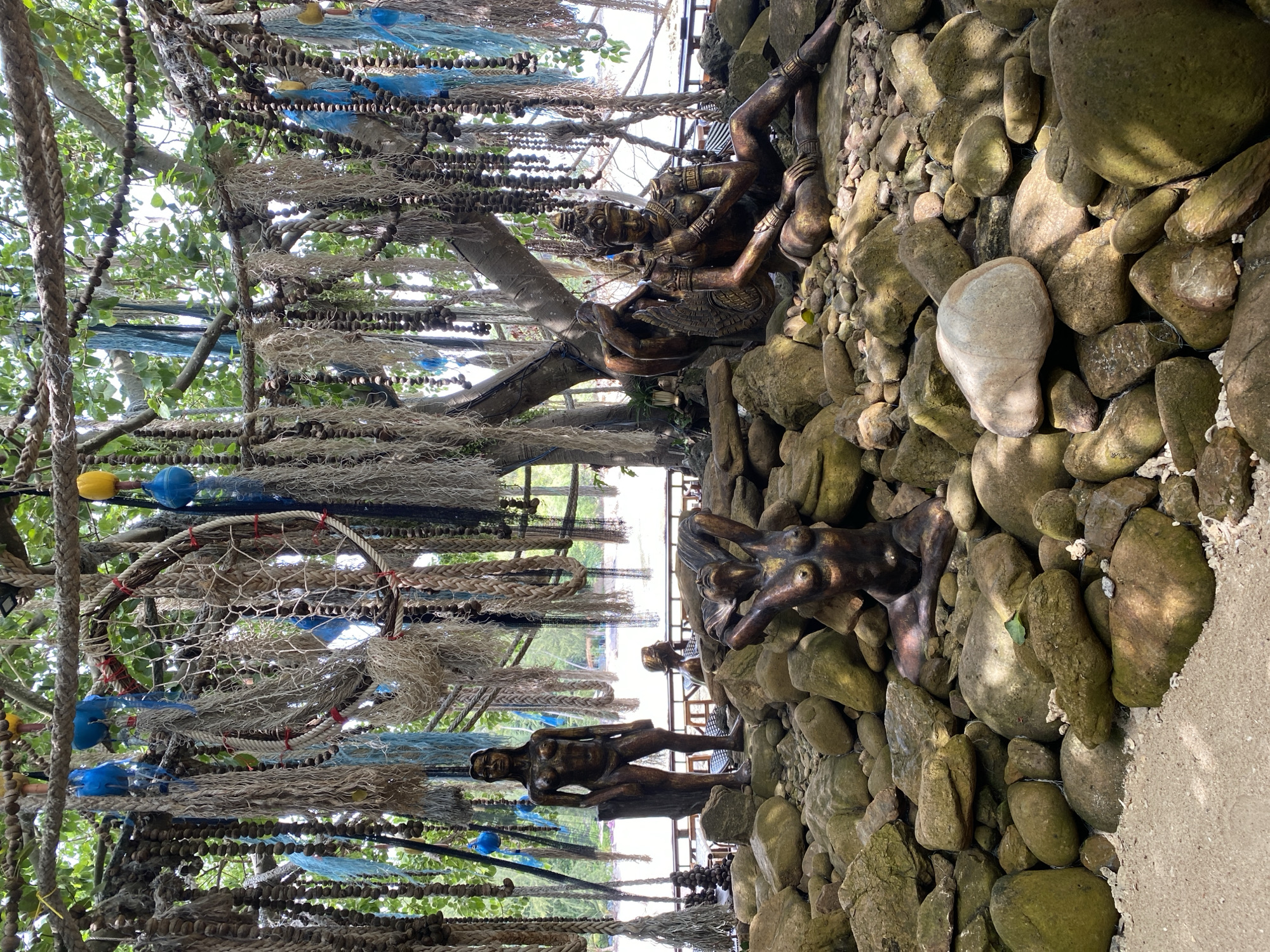 The image depicts an outdoor scene under a tree decorated with numerous ropes, nets, and blue and yellow buoys. In the foreground, scattered large, moss-covered rocks form a naturalistic base for several bronze sculptures. Three distinct sculptures are visible: a standing female figure on the left, a seated figure with wings and intricate jewelry in the center right, and a reclining female figure with her arms behind her head in the lower center. The background shows a body of water, possibly the sea, with a distant shoreline that includes structures and vegetation. The overall setting suggests a tropical or coastal environment, likely in a resort or public space in ตำบลเกาะพะงัน, Thailand, given the presence of decorative elements and sculptures commonly found in such locations. The lighting indicates it is daytime, with shadows suggesting the sun is overhead or slightly to the side.