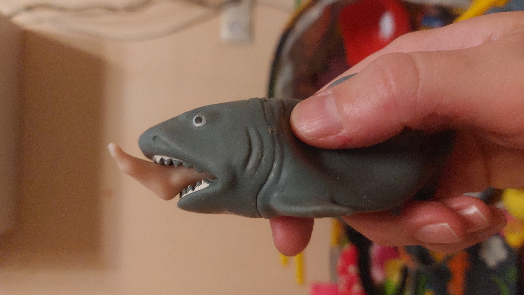 A person's hand holds a gray, rubber toy shark. The shark's mouth is open, and a finger is inserted inside, appearing to be bitten. The background is blurry but appears to be an indoor setting. It is impossible to determine whether the picture was taken in Novosibirsk, Russia without additional information.