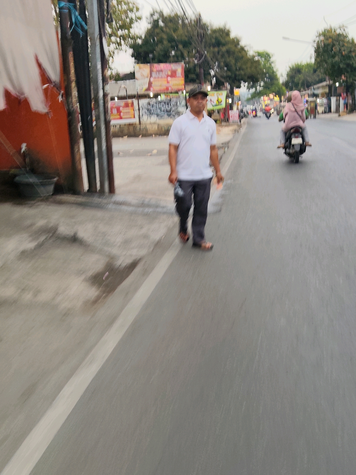 A man wearing a white polo shirt, dark pants, a dark baseball cap, and sandals walks on the right side of a road in Purwakarta, Indonesia. He is carrying a dark object in his right hand. A person on a motorcycle wearing a pink hijab is riding in the same direction on the road. Buildings with signs and trees line the street. The sky is overcast. The road appears wet, suggesting recent rain. The image is blurry due to motion.