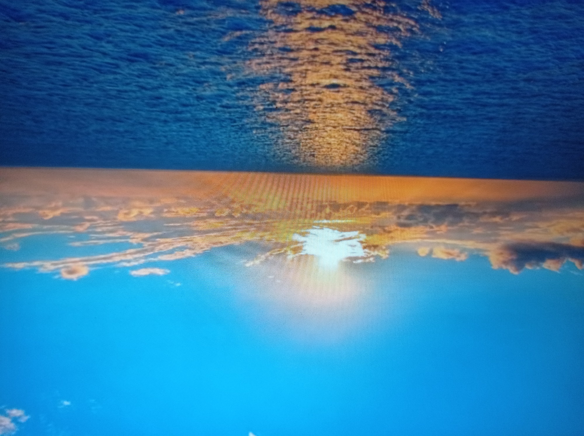 The image displays a wide expanse of ocean under a bright blue sky with scattered clouds. The sun is prominently featured, casting a strong reflection on the water's surface, creating a shimmering path of light. The horizon line is clearly visible, separating the deep blue of the sea from the sky and clouds. The clouds are tinged with warm, golden hues, suggesting either sunrise or sunset. The image is framed from a perspective that implies being at sea, likely from a boat or ship, given the wake visible on the right side of the ocean. There are no people, buildings, or any other discernible man-made objects present in the scene. The environment depicted is purely natural, focusing on the vastness of the ocean and sky. The location is not specified as being in the city of Mukachevo, Ukraine, as this is a natural seascape and Mukachevo is a landlocked city.