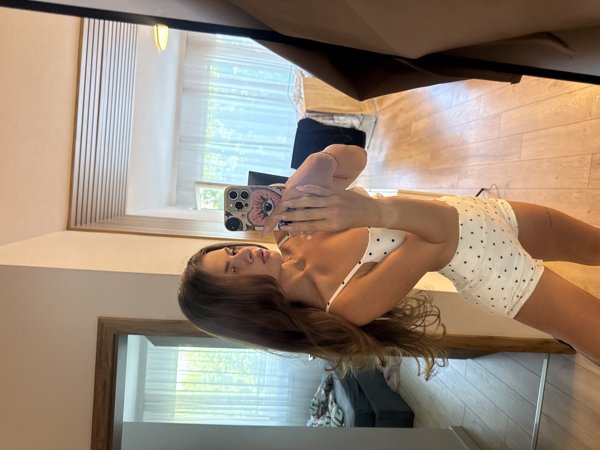 The image shows a young woman taking a selfie in what appears to be a bedroom or dressing area. She is wearing a white polka dot camisole and shorts. She is holding a phone with a decorated case, which is reflecting light. The background shows a mirror reflecting more of the room, including a bed with a patterned duvet, a window with sheer curtains, and a wooden floor. The overall impression is that of a casual, personal moment captured in a domestic setting. This location is not identifiable as being in Plovdiv, Bulgaria.