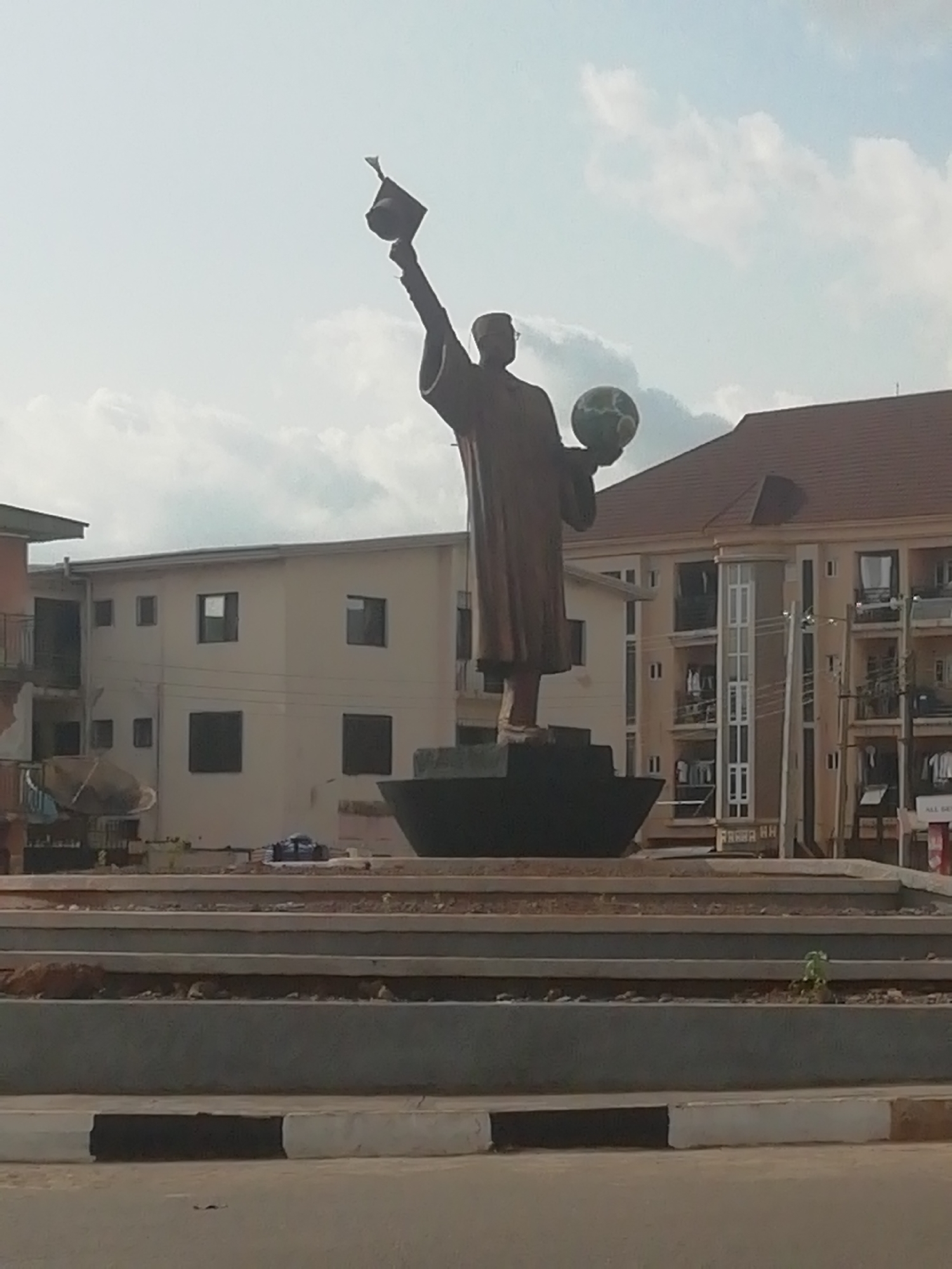 The image shows a bronze statue of a man on a traffic circle. The man is wearing a robe and holding a graduation cap in his right hand and a globe in his left hand. The statue is located in Enugu, Nigeria. The background of the image shows several buildings.