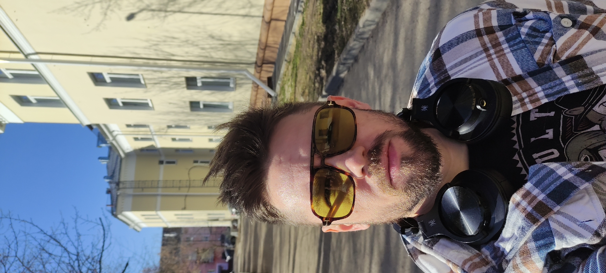 The image shows a man standing outside. He has dark hair and a beard and is wearing sunglasses, a black T-shirt, and a plaid shirt. He is wearing headphones around his neck. The background shows a bright blue sky, buildings with multiple windows, bare tree branches, and a paved area. The buildings and overall layout suggest the image was taken in an urban area. The city is Fryazino, Russia.