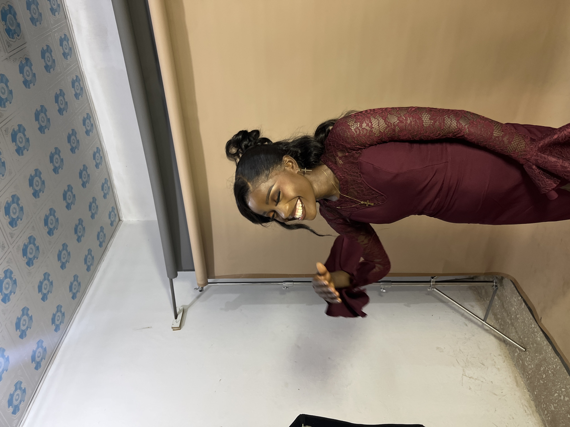 In Gura Top, Nigeria, a young Black woman with her hair styled in a high bun and loose waves stands in a studio setting. She is wearing a deep red, long-sleeved dress with lace detailing on the sleeves and neckline. She is smiling with her eyes closed and her left hand is raised, palm facing forward. A brown backdrop is visible behind her, and a white wall with a patterned ceiling is to her left. A photography stand is present in the foreground.