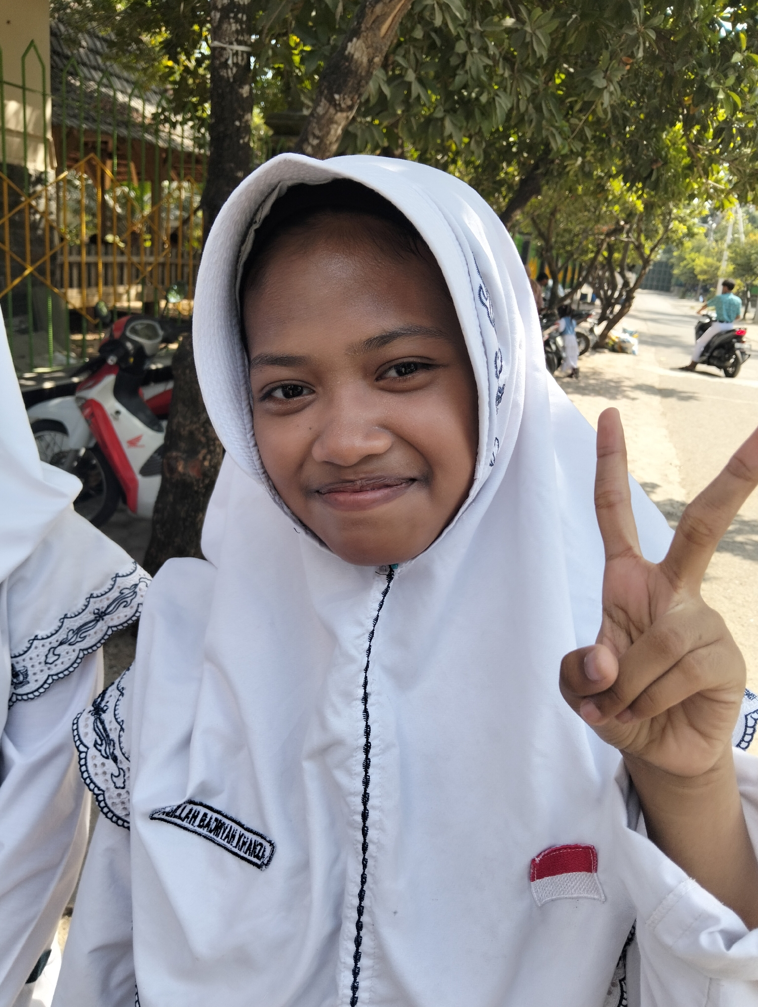 A young Indonesian girl wearing a white hijab and uniform smiles at the camera and holds up two fingers in a peace sign. She is standing outdoors in what appears to be a schoolyard in Paciran, Indonesia. Behind her, a tree casts a shadow, and a motorcycle is parked. In the background, a street is visible with more motorcycles and people. The girl's uniform has an embroidered patch with text and a small Indonesian flag. The sunlight suggests it is daytime.