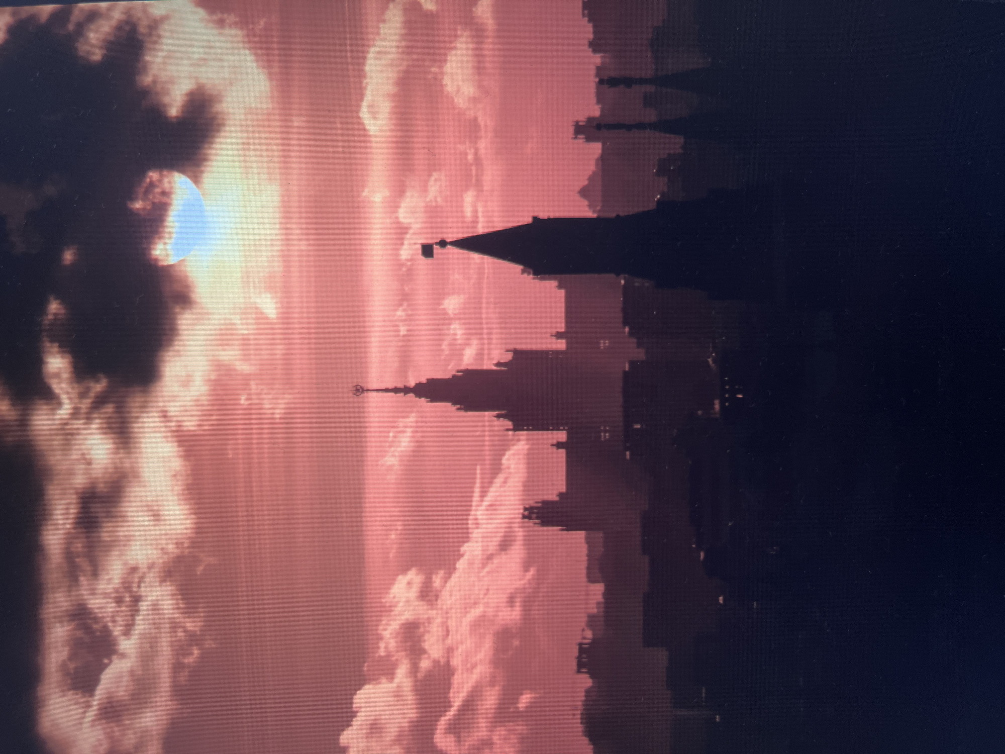The image depicts a silhouetted cityscape against a dramatic sky with a crimson hue. In the foreground, the dark outlines of various buildings rise against the vibrant sky. A prominent spire with a flag on top is visible, along with other pointed structures. Behind these, a larger, more imposing building with a central spire dominates the middle ground. The sky is filled with large, textured clouds, with a portion of a bright, circular object, likely the sun, peeking through the clouds, casting a pale blue and yellow light. The overall impression is one of a dramatic sunset or sunrise over an urban landscape, with the buildings appearing as dark silhouettes against the intensely colored sky. The location is identified as the city of Khimki, Russia.