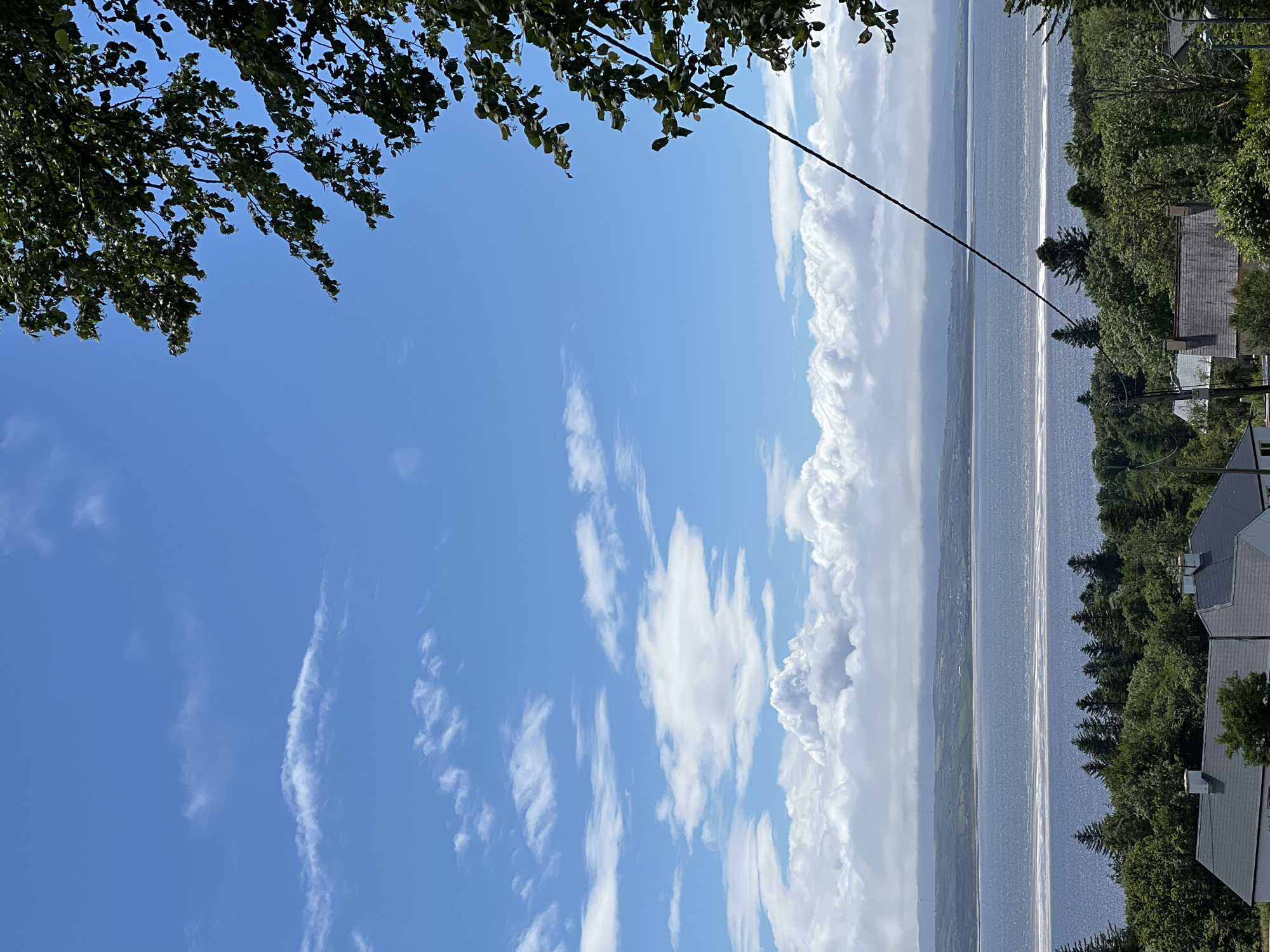 This is a vertical, eye-level, outdoor, daytime shot of a scenic view overlooking a body of water, likely a bay or large river, from a slightly elevated position. The scene is framed by dark green tree branches in the upper left corner. The sky is a vibrant blue with scattered white clouds, some of which are large and puffy, while others are wispy streaks. A thin, dark line, possibly a wire or string, extends diagonally from the top center towards the lower right.
The water stretches horizontally across the middle of the frame, reflecting the sunlight with a shimmery, textured appearance. In the distance, faint landforms are visible under a hazy sky. To the right of the water, a dense line of dark green trees borders the edge of the land. Several houses with dark grey roofs are nestled among the trees, suggesting a residential area. The architecture of the houses appears to be traditional.
The overall impression is one of a peaceful, natural landscape combined with human habitation. The time of day appears to be late morning or early afternoon, given the bright sunlight and clear blue sky. The weather seems pleasant and sunny. While no people are visible, the presence of houses indicates human activity in the area. There is no visible text in the image. The location context places this scene in Mountcharles, Ireland.