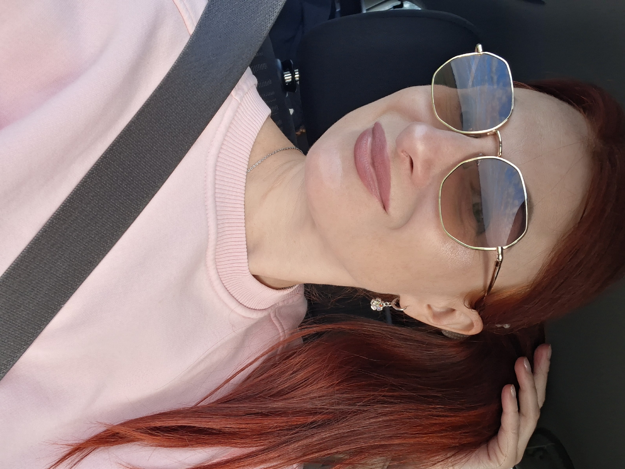A woman with red hair is in the driver's seat of a car, taking a selfie. She is wearing sunglasses with gold frames and pink lipstick. A seatbelt is visible across her chest. The car interior is dark, suggesting it is daytime. The woman's hand is in her hair, and she is smiling. The background shows a blurred landscape reflected in her sunglasses, which appears to be sky and land.