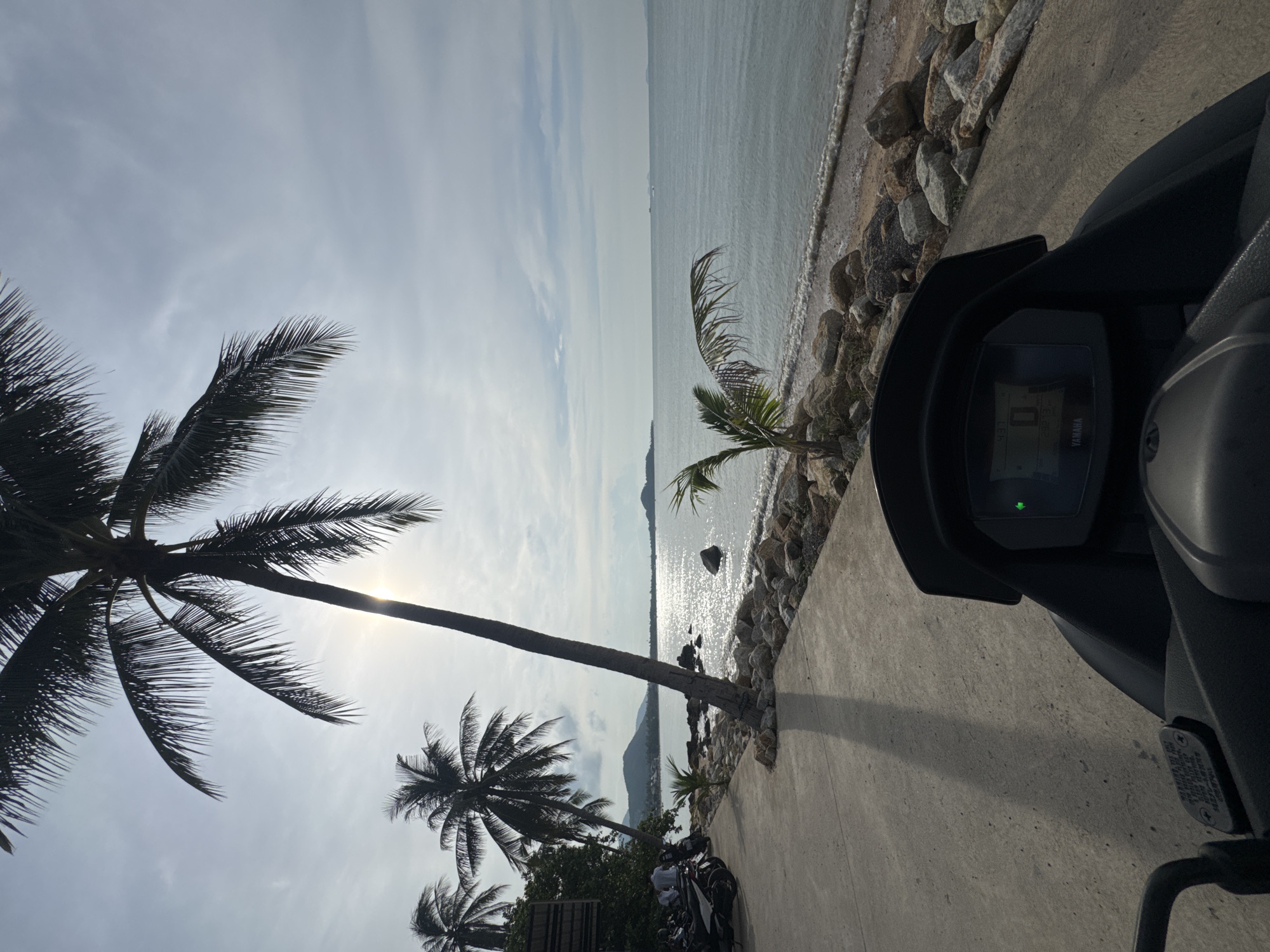The image depicts a perspective from a motorcycle parked on a paved walkway overlooking a tropical beach scene. Tall, silhouetted palm trees dominate the left side of the frame, with their fronds extending into a hazy sky. The sun is partially obscured by a palm tree, casting a bright glare on the water and creating a shimmering effect on the ocean's surface. The sea is calm with gentle waves lapping against a rocky shoreline. In the distance, the coastline is lined with more palm trees and appears to have some buildings or structures. Several parked motorcycles are visible on the left side of the walkway, suggesting a stop or resting point. The walkway itself is made of concrete and shows a distinct shadow from the prominent palm tree. The digital display of the motorcycle's dashboard is visible in the foreground, showing some icons and text, including the Yamaha brand. The overall scene evokes a sense of a serene tropical environment, likely in the Ko Samui District of Thailand, given the presence of palm trees and the coastal setting.