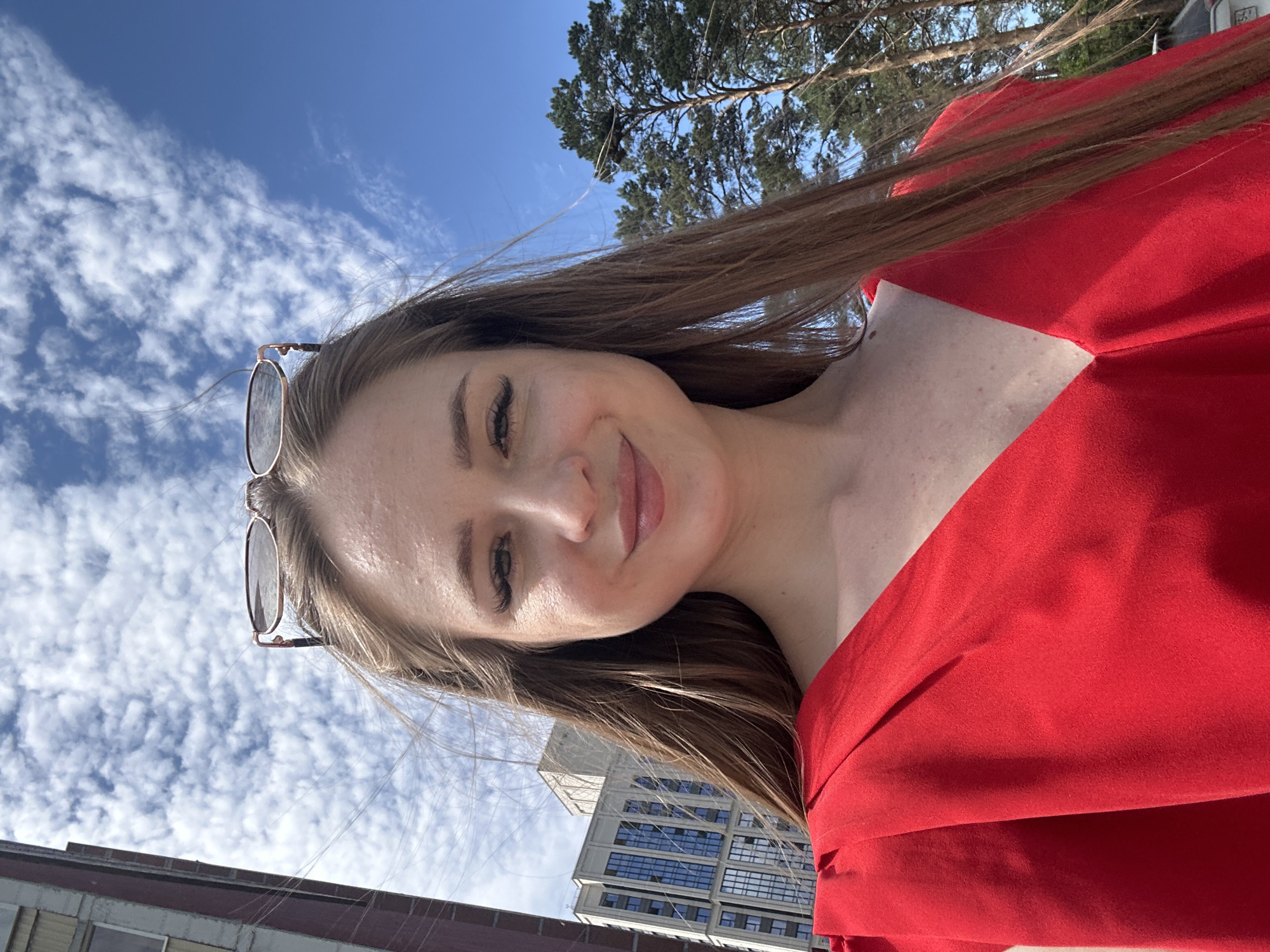 An outdoor, waist-up, medium-shot photograph features a woman with long, brown hair and brown eyes, wearing a vibrant red dress and rose-gold rimmed glasses on top of her head. She is smiling broadly, looking directly at the camera. Her skin is fair with some visible freckles and small blemishes on her chest. The background is a bright blue sky filled with scattered white clouds. To the left of the woman, a modern, multi-story building with blue-tinted windows is visible, and to her right, tall pine trees stand against the sky. The lighting suggests a sunny day, casting subtle shadows and highlighting the texture of her dress. The image appears to be a selfie.