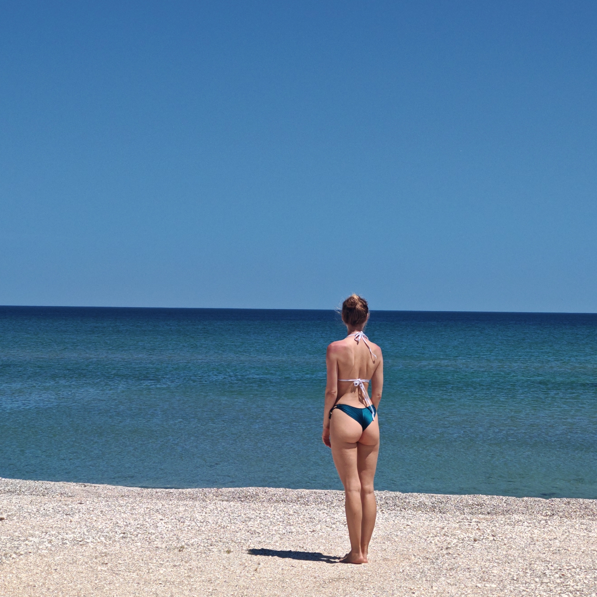 A woman stands on a beach facing the ocean. She wears a two-piece bathing suit. The beach appears to be composed of light-colored pebbles or shells. The sea is calm and blue, meeting a clear blue sky. The location is Unknown, Unknown.