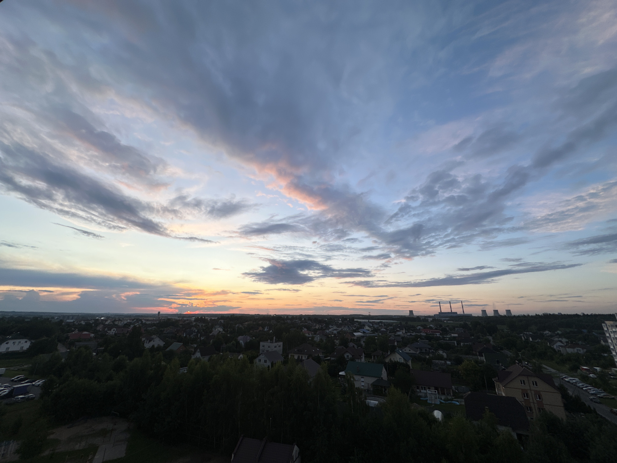This is a wide-angle photograph of a residential area at sunset, likely within or near Шчомысліцкі сельскі Савет, Belarus. The sky dominates the upper two-thirds of the image, featuring a dramatic sunset with streaks of pink, orange, and blue clouds against a backdrop of twilight blue. Below the sky, a sprawling landscape of houses, trees, and distant industrial structures is visible. The houses are of various sizes and architectural styles, many with dark roofs. Dense green trees are prevalent throughout the scene, forming a dark, almost silhouetted border in the foreground and scattered throughout the residential areas. In the mid-ground, a cluster of industrial cooling towers or chimneys can be seen against the horizon, emitting plumes of steam or smoke. A road lined with parked cars is partially visible in the mid-right of the image. The overall impression is of a suburban or semi-rural environment at the end of the day. There are no people visible in the image.