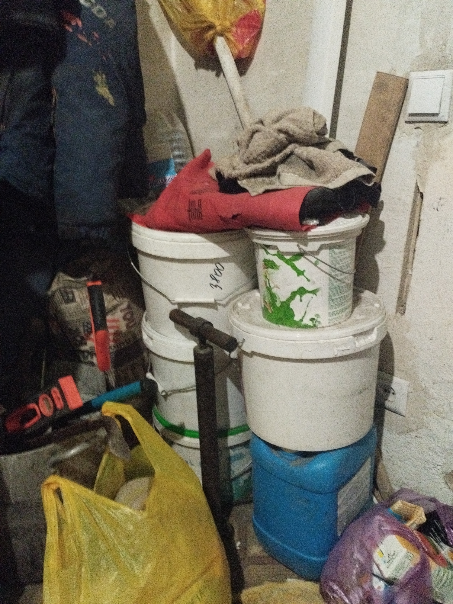 The image depicts a cluttered storage area, likely a workshop or shed, with various construction and household items. In the foreground, a bright yellow plastic bag is open, containing what appears to be light-colored material. To the right of the bag is a blue plastic container, topped by two white buckets stacked on top of each other. The top bucket has a red lid with some sort of fabric draped over it, and the lower bucket has green splatters on its side. A metal pipe structure is visible in front of the buckets, extending downwards. To the left of the yellow bag, an orange and black tool, possibly a drill or a saw, lies on a dark surface. Behind the yellow bag, a dark blue garment, possibly a jacket or overalls, hangs on the left side of the frame. The background consists of a light-colored, textured wall with a light switch and an electrical outlet visible on the right side. A piece of wood leans against the wall behind the buckets. A yellow plastic bag is partially visible at the top of the frame. The overall impression is one of disarray and storage of various materials. The scene is located in the city of Sarykol, Kazakhstan.
