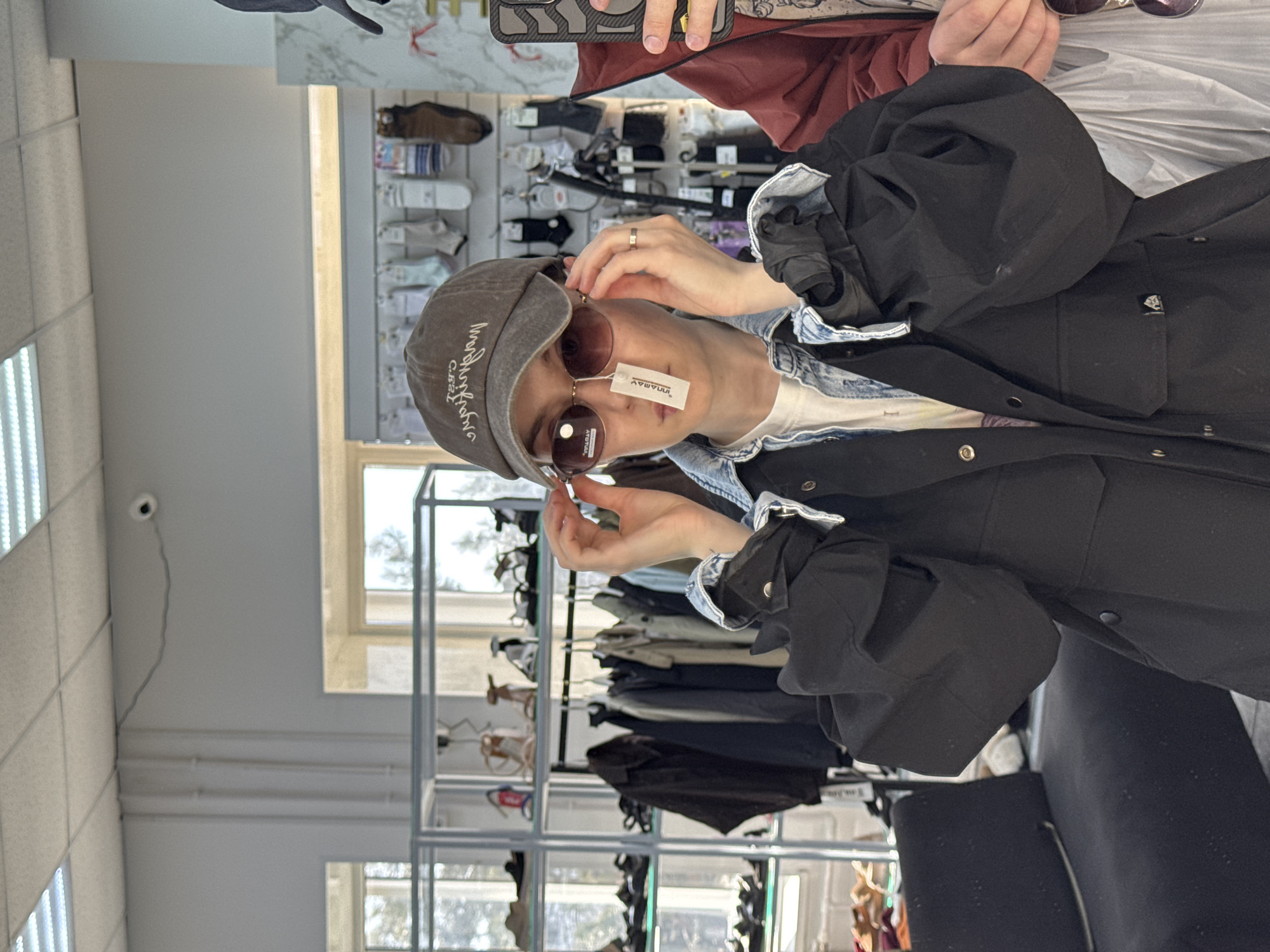 The photo shows a person in a store, likely in Chaikovsky, Russia. The person is wearing a gray cap with Cyrillic text, sunglasses with a price tag attached, a denim shirt, and a black jacket with large sleeves. They are holding the sunglasses up, seemingly trying them on. Behind them is a metal rack with various clothing items and shoes. In the background, there is a window with daylight coming through. A security camera is visible on the ceiling. Another person, holding a smartphone, is partially visible at the top of the frame.