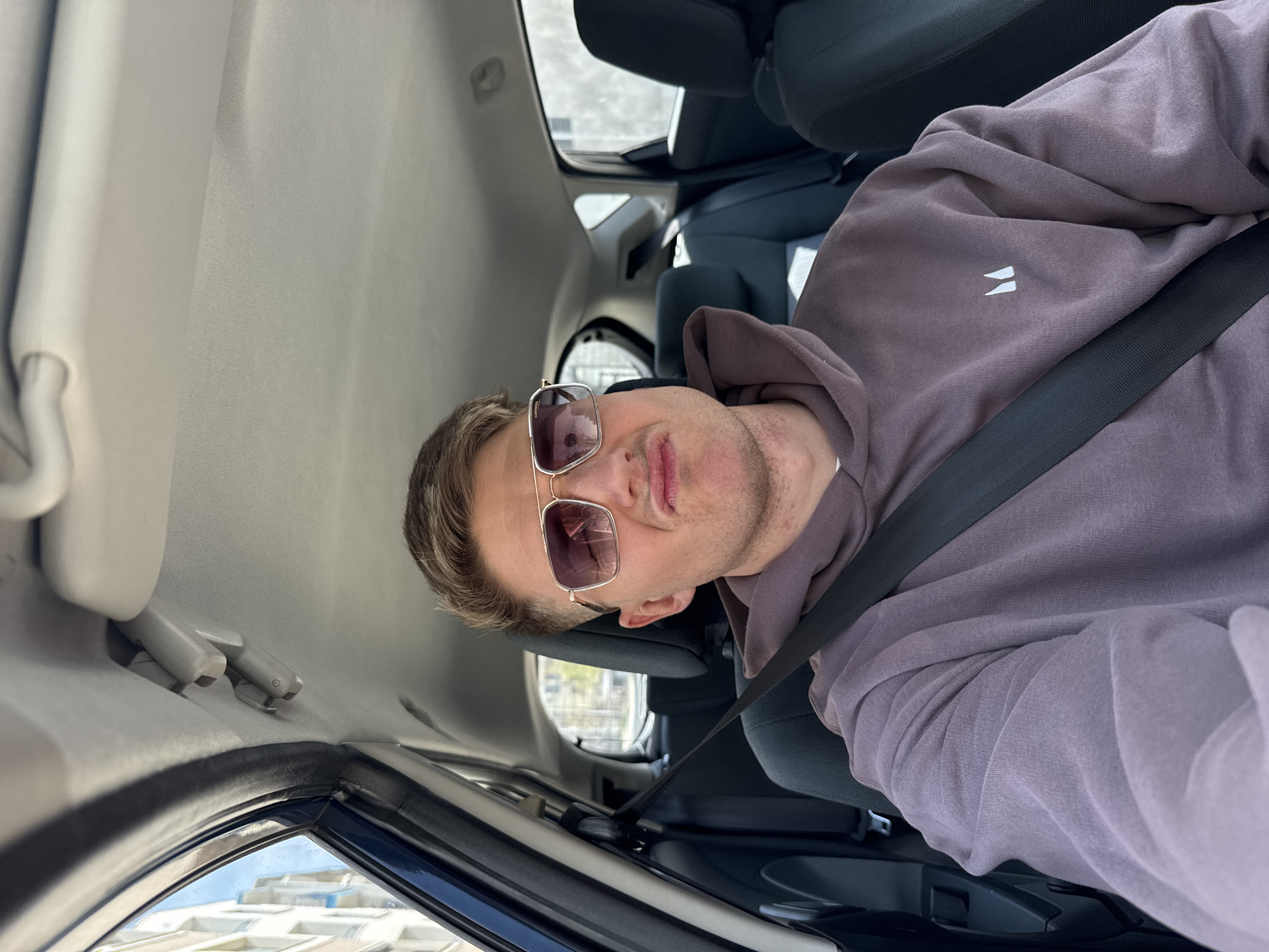 A male is seated in the back seat of a car wearing sunglasses, a lavender hooded sweatshirt, and a seatbelt. The car's interior is visible, showing the headliner and window frame. Buildings are visible outside the car window. Based on the visible architecture and urban layout, the image likely depicts a location in Burgas, Bulgaria.