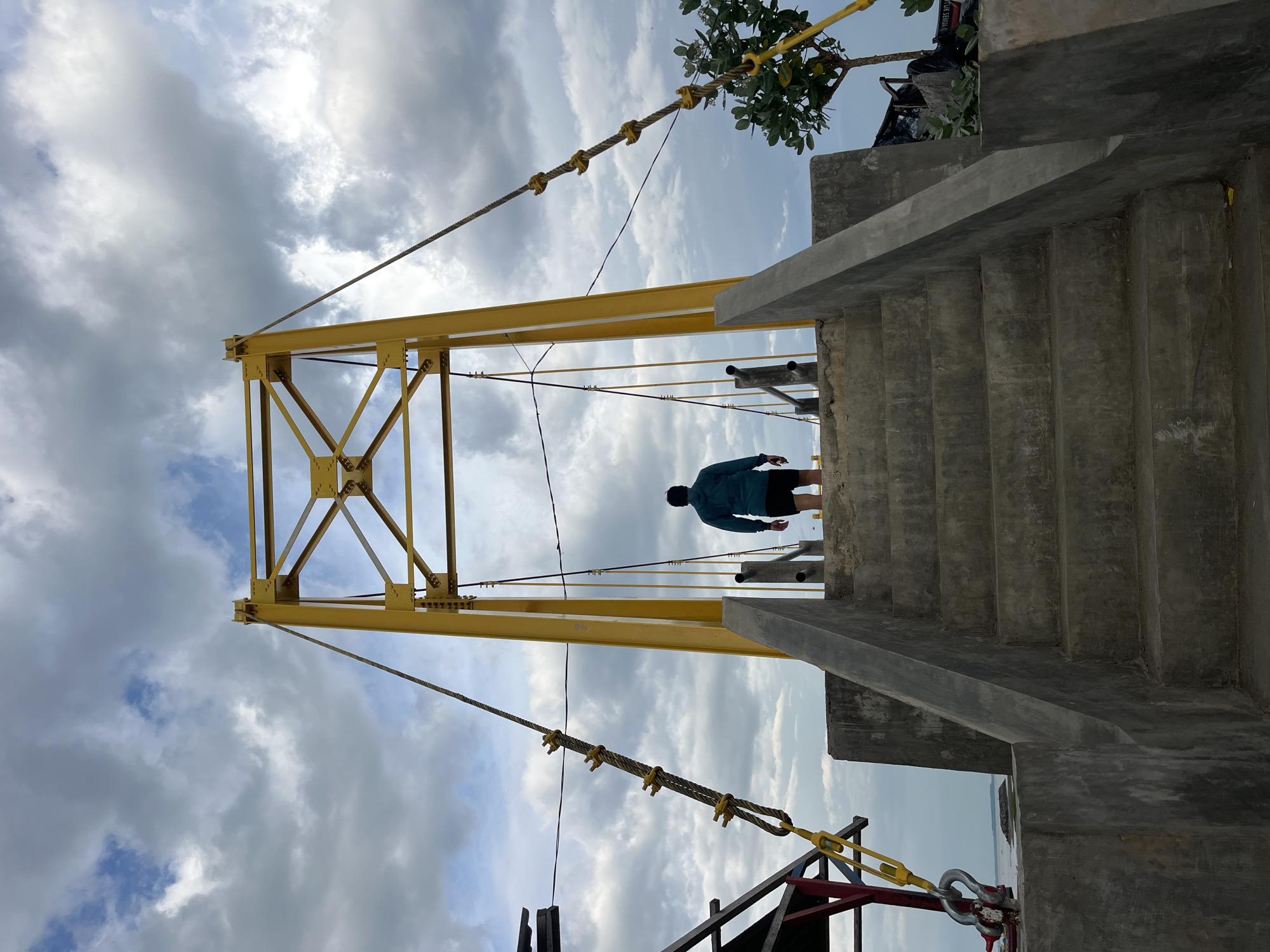 A low-angle shot, slightly skewed, shows a yellow structural tower connected to a bridge over a body of water near the city of Lhokseumawe, Indonesia. A person is standing on the bridge. The sky is visible behind the tower, and partially cloudy. Concrete steps lead up to the bridge on the right side of the frame. A tree is visible at the top of the stairs.